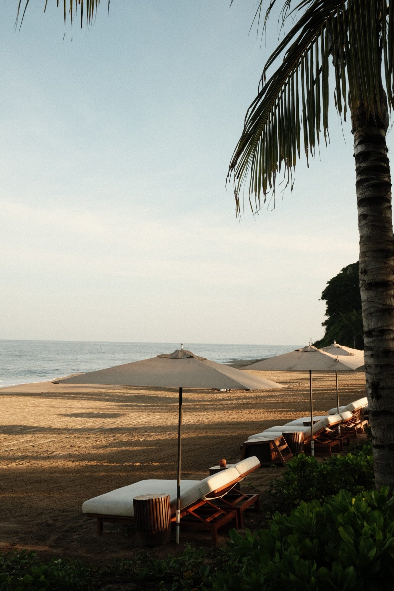 Rosewood Mandarina, Mexico — Teak loungers and canvas umbrellas on sand beach at dusk, palm fronds overhead, calm ocean horizon beyond.