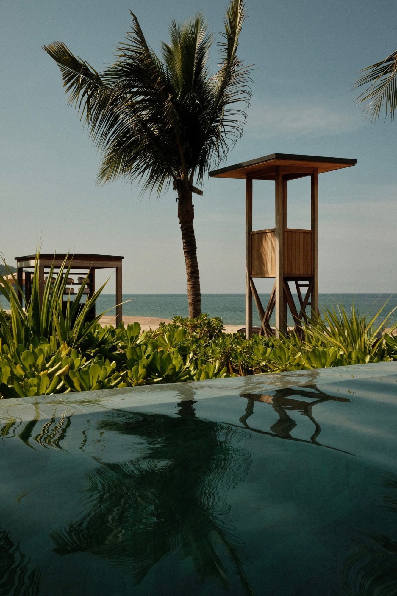 Rosewood Mandarina, Mexico — Wooden lifeguard tower and palm tree overlooking calm ocean, reflecting in wet sand with coastal vegetation and clear sky.