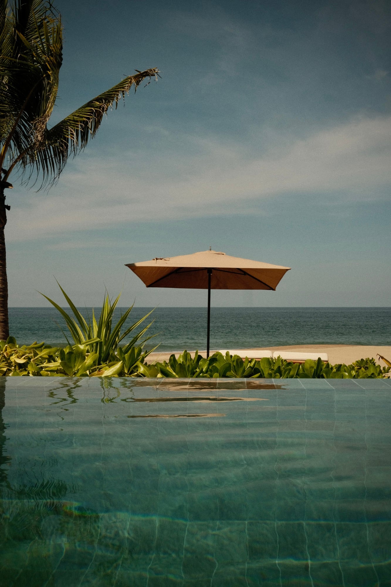 Rosewood Mandarina, Mexico — Beige rectangular umbrella beside infinity pool overlooking calm ocean, palm fronds and coastal vegetation in foreground, midday light.