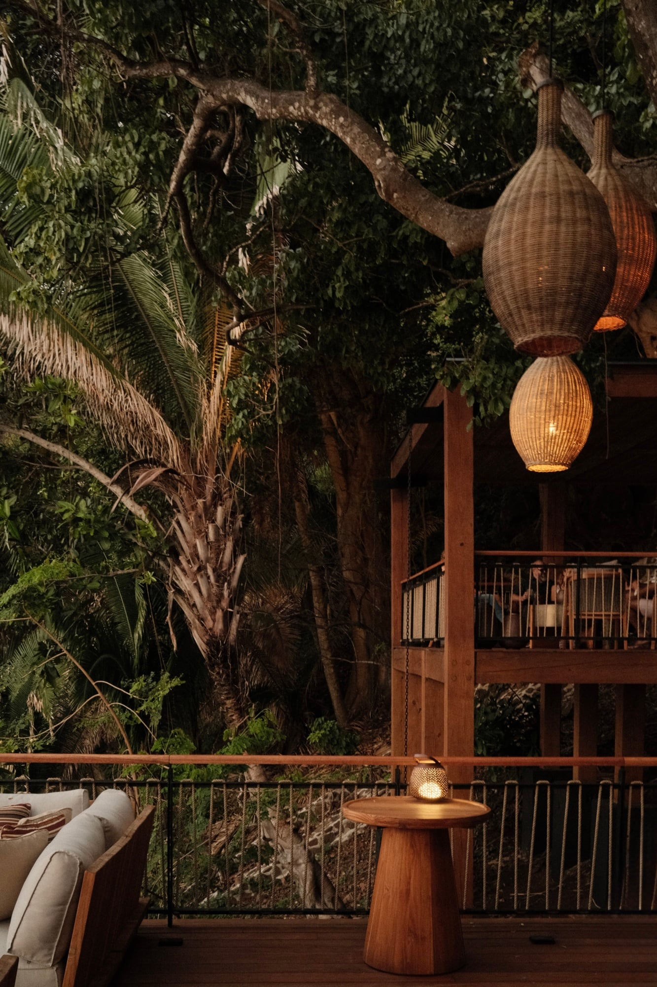 Barra Peñasco, Mexico — Woven pendant lights hang above wooden deck overlooking jungle canopy at dusk, rattan furnishings and metal railings define contemporary tropical retreat.