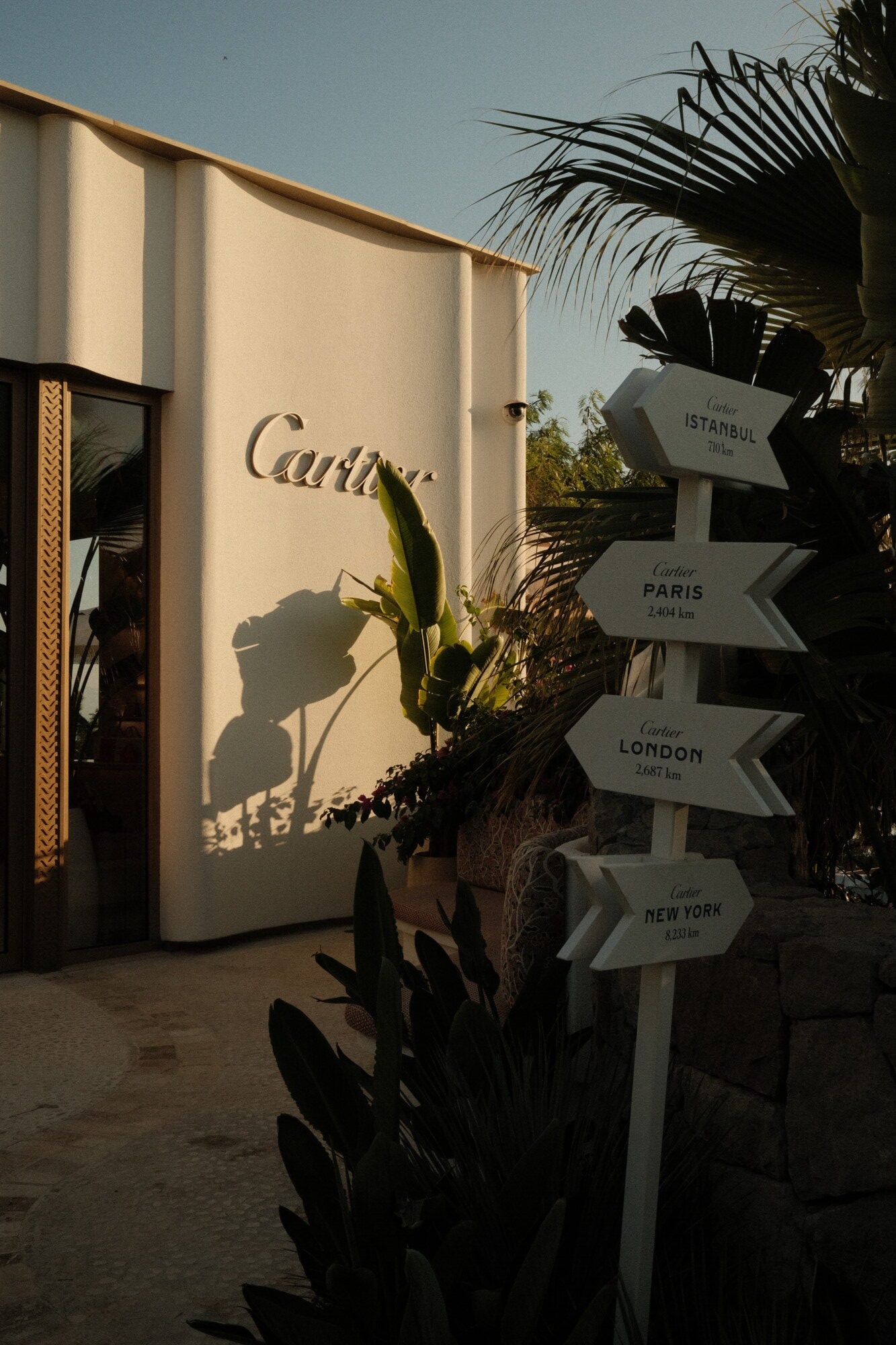 Mandarin Oriental Bodrum, Turkey — Cartier boutique exterior with directional signage to Paris, London, Istanbul, white stone facade catching golden afternoon light, tropical plants casting shadows.
