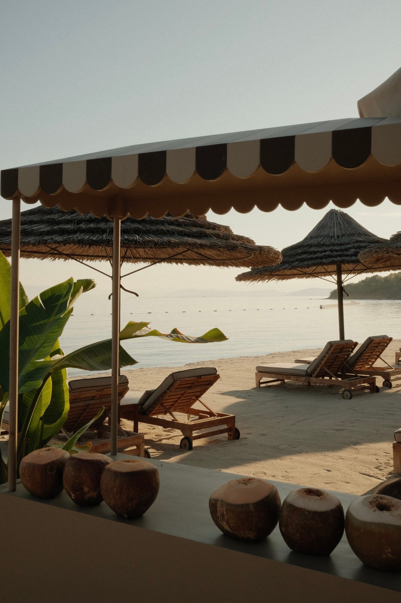Mandarin Oriental Bodrum, Turkey — Striped canvas awning and thatched umbrellas shade wooden loungers on sandy beach overlooking calm bay at dusk.