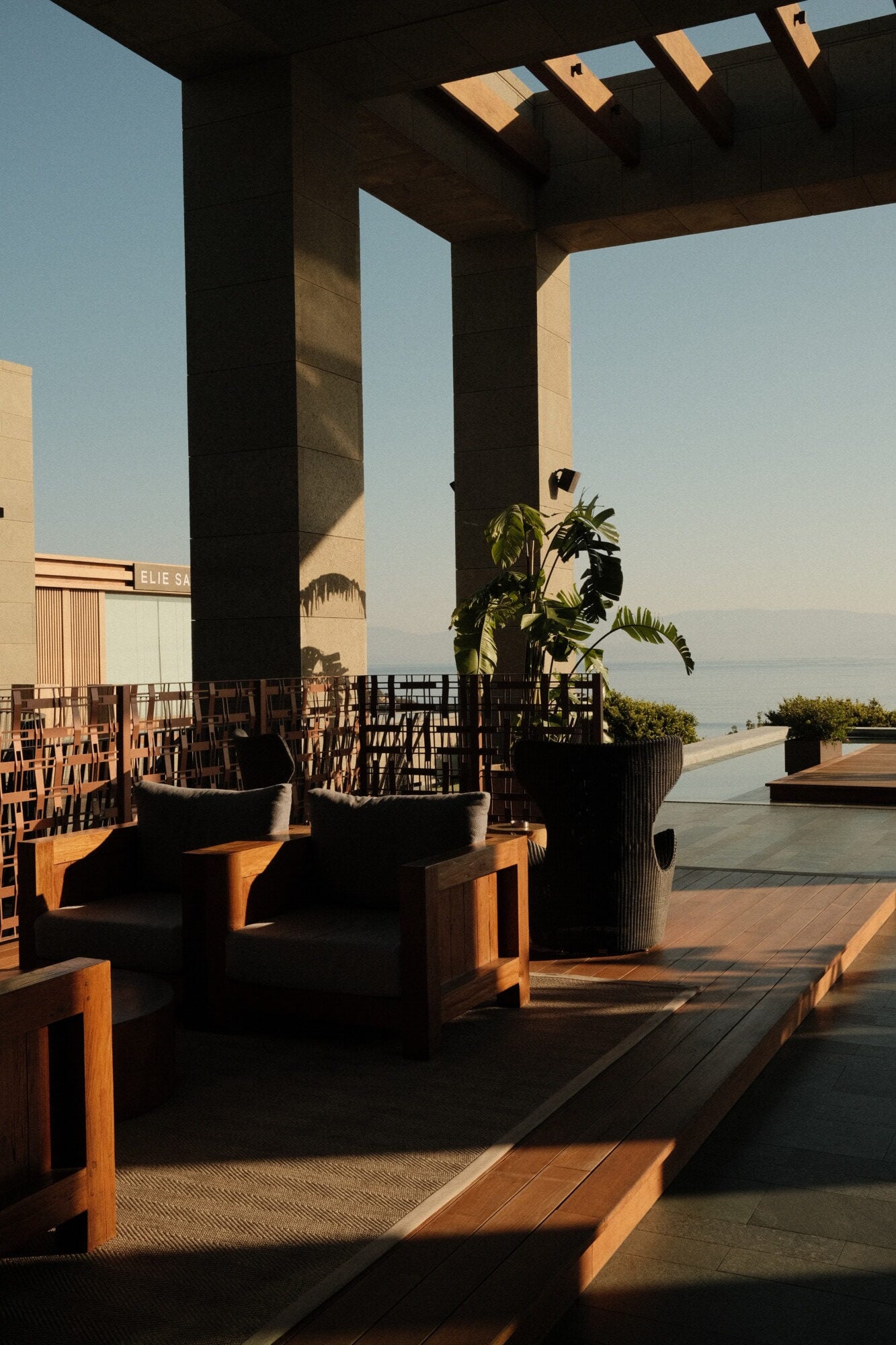 Mandarin Oriental Bodrum, Turkey — Modern waterfront terrace with dark pergola, wooden seating, potted plants, and Mediterranean Sea views at golden hour.
