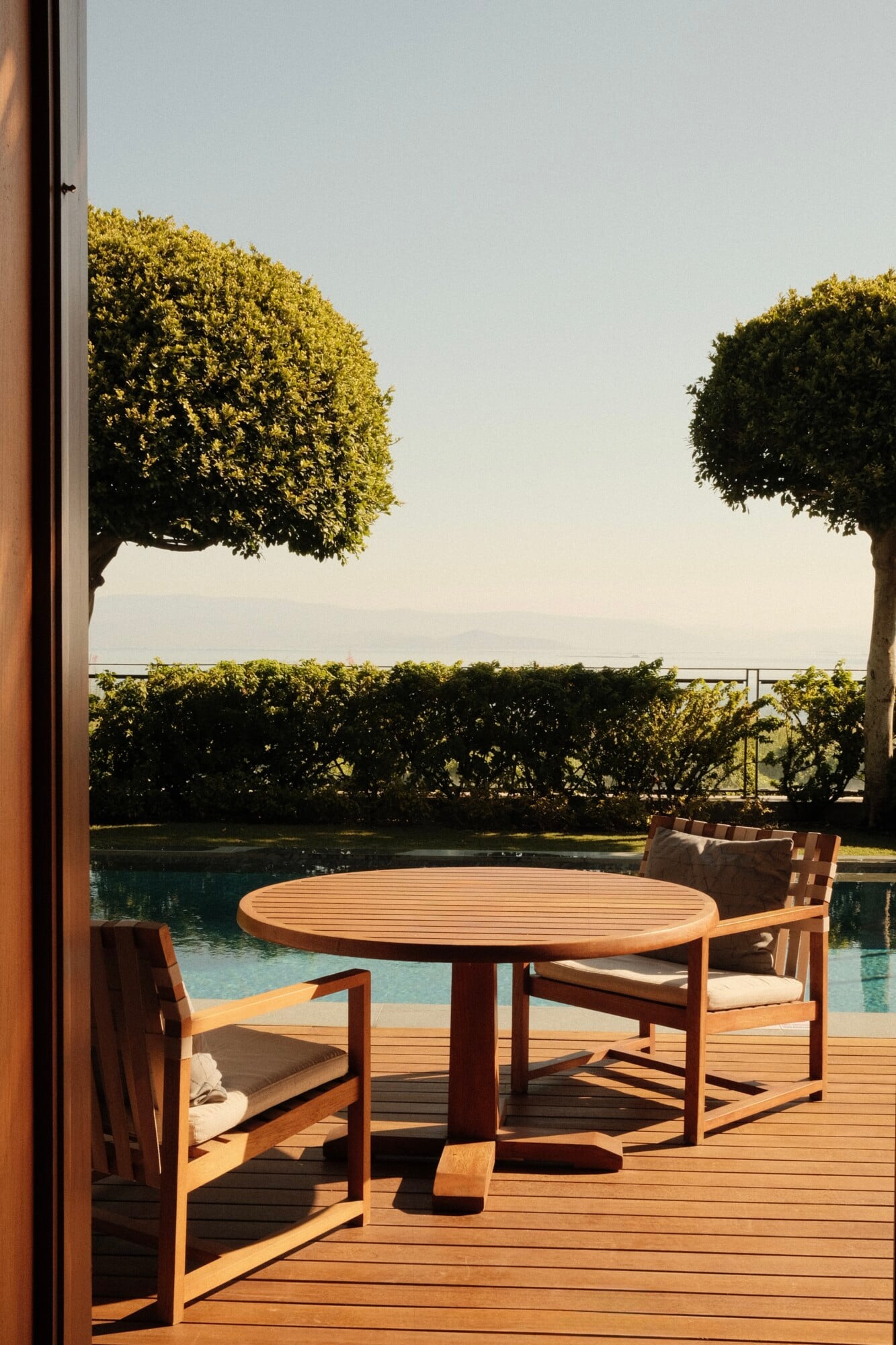 Wooden deck with round table and chairs overlooking a pool and bay, framed by sculpted hedges in golden afternoon light.
