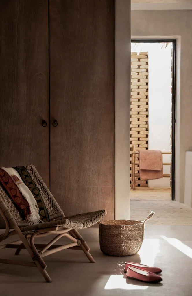 The Rooster, Europe — Dark wood wardrobe, woven chair with layered textiles, natural light, minimalist bedroom interior with brick detail visible through doorway.