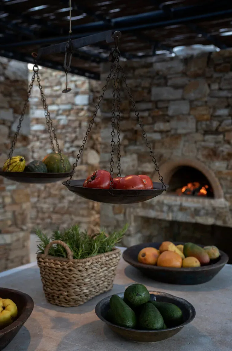 The Rooster, Europe — Hanging cast-iron baskets of fresh produce beside stone fireplace with rosemary herb pot and arranged fruits on table.