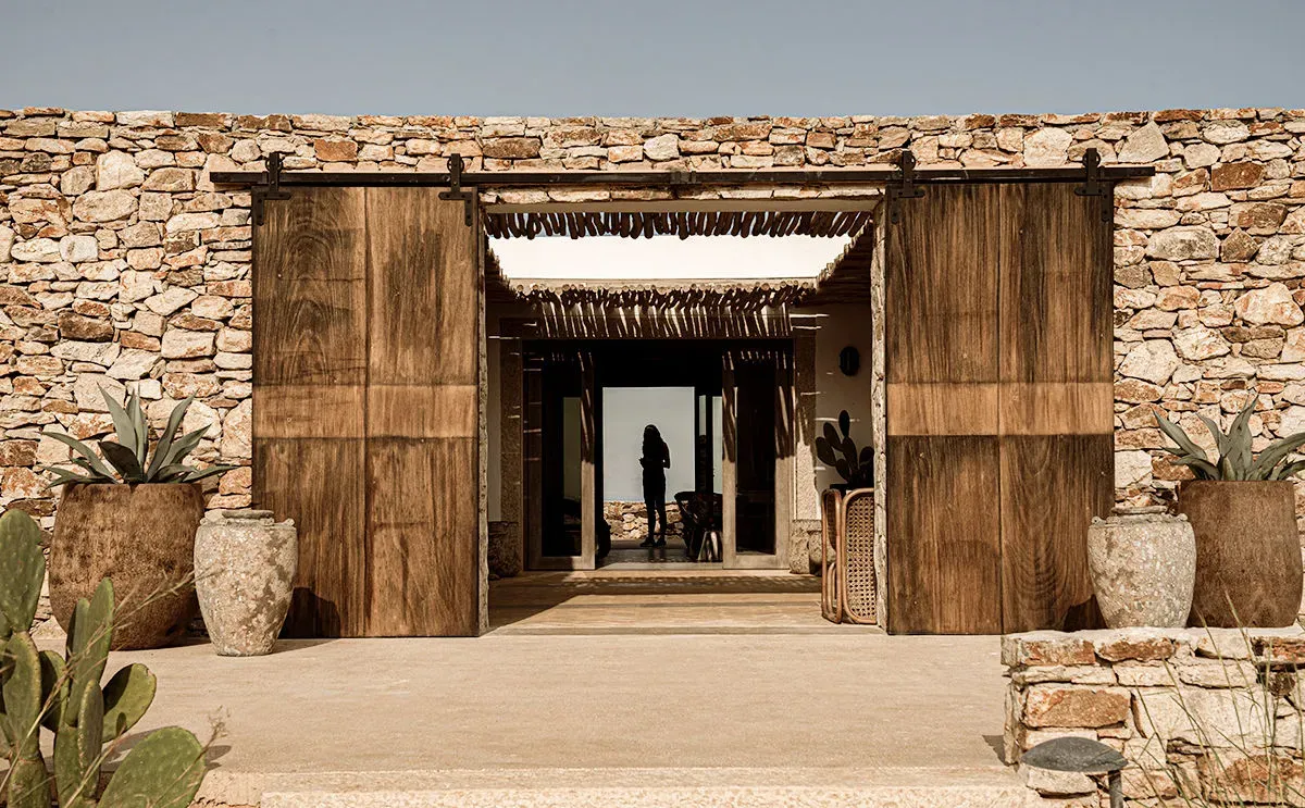 The Rooster, Europe — Stone-walled entrance with dark wooden sliding doors, terra-cotta planters, and figure silhouetted in bright passage beyond.