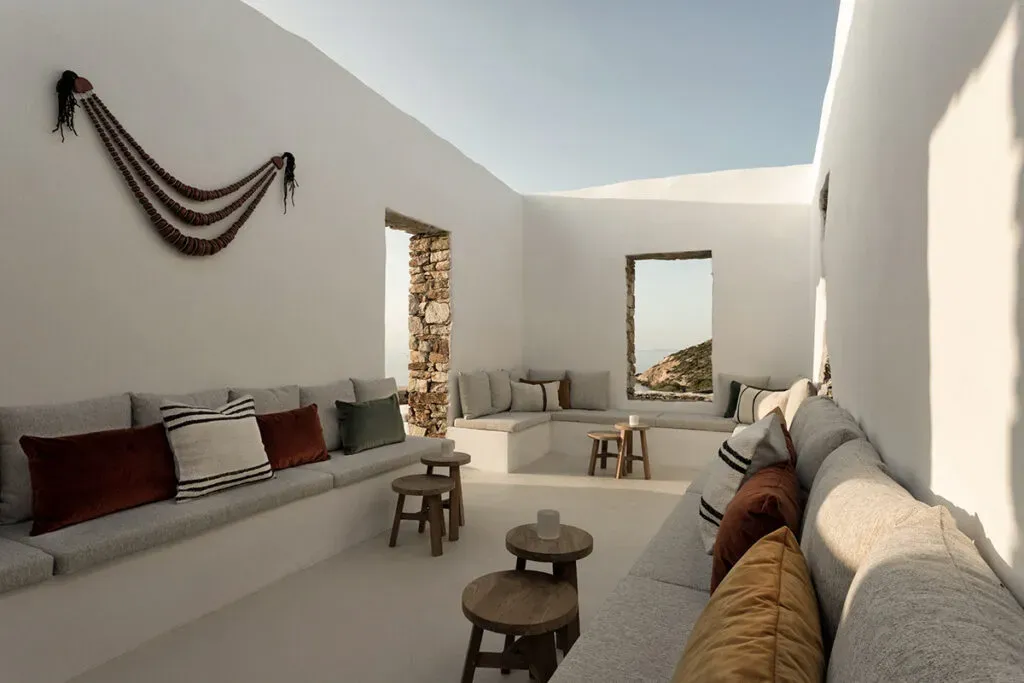 The Rooster, Europe — Sunlit minimalist lounge with stacked linen seating, stone archways, wooden stools, layered textiles, and sculptural wall hanging.
