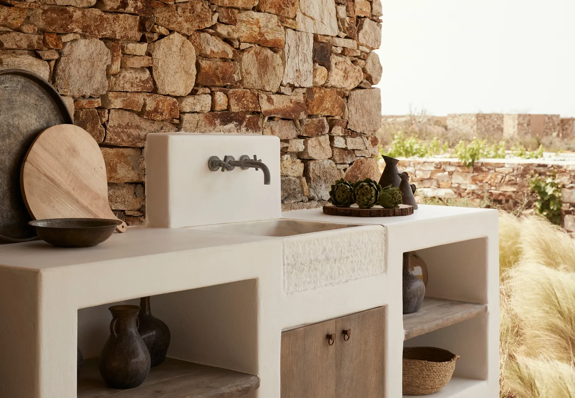 The Rooster, Europe — Outdoor kitchen sink with white plaster counter, aged brick wall, dark brass faucet, wooden accessories, and terracotta vessels.