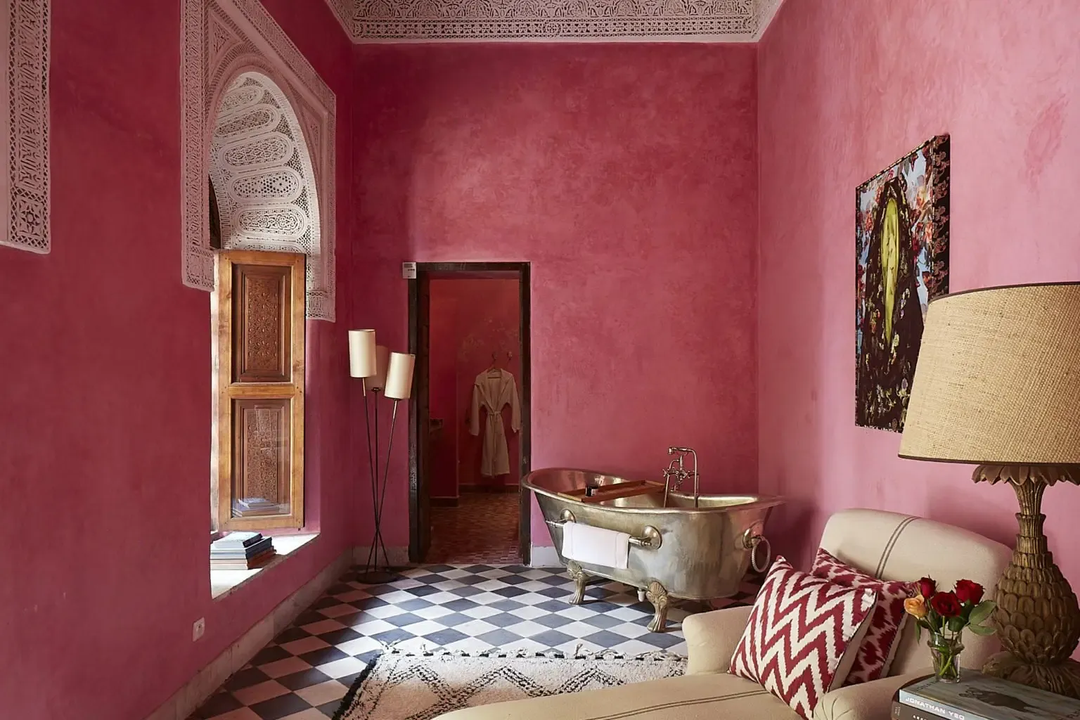 El Fenn, Marrakech, Morocco — Magenta-walled bathroom with zellige tilework, arched carved doorway, brass bathtub, and black-and-white checkered floor.