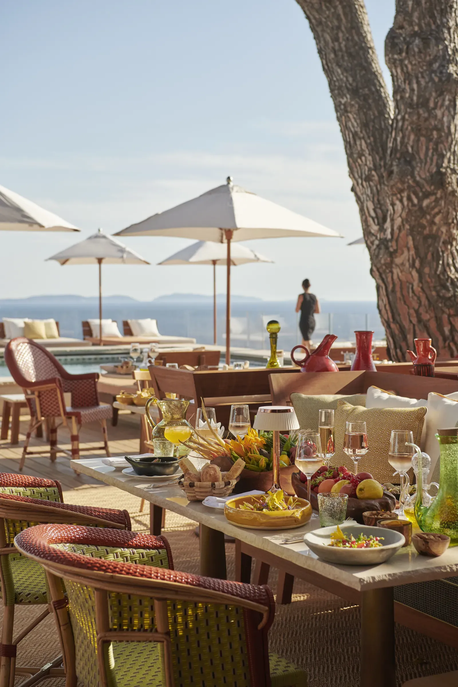 Lily of the Valley, Europe — Seaside terrace with linen umbrellas, stone dining table, coral chairs, olive oil service, sea view, and mature tree trunk in afternoon light.