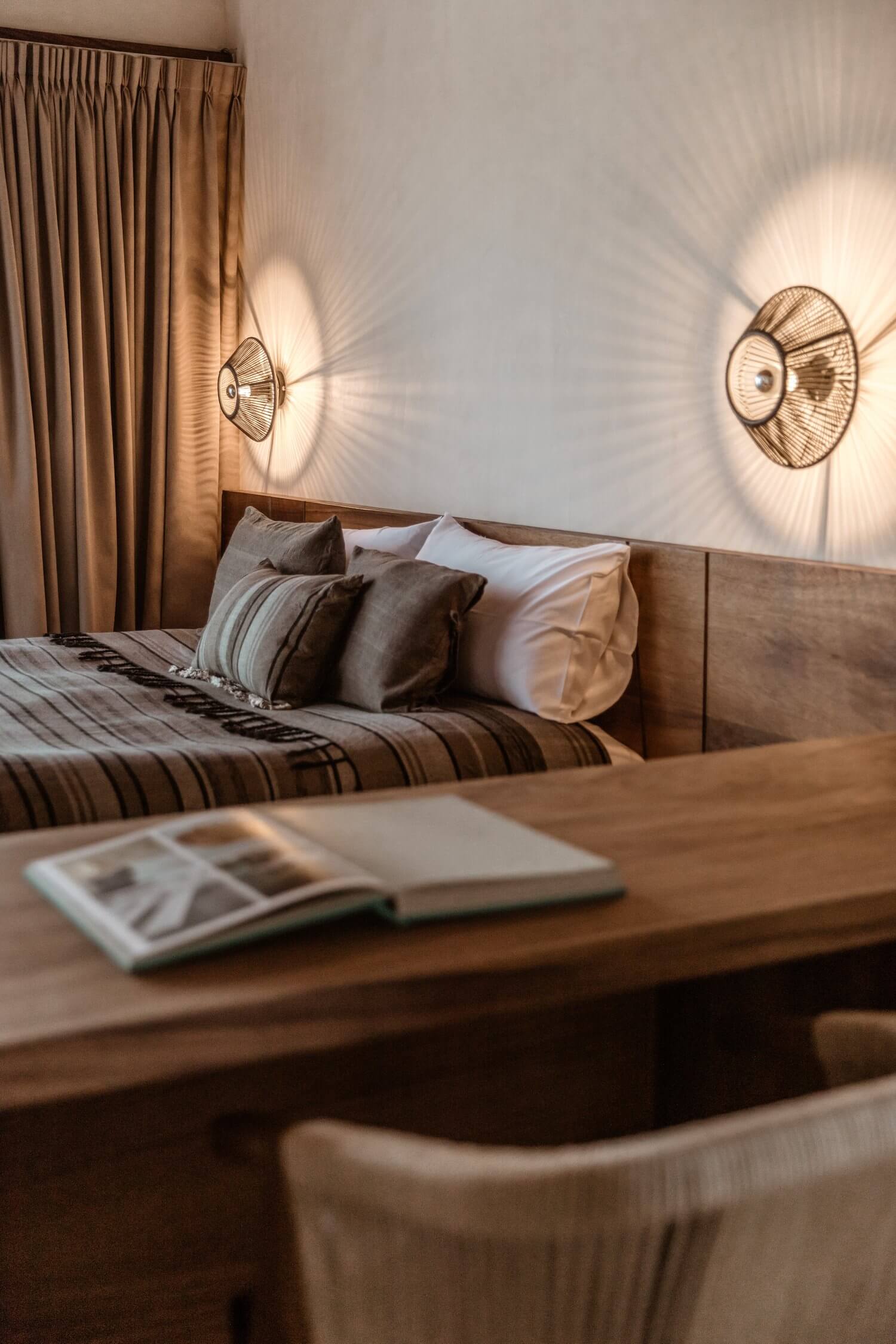 Hotelito MUSA, Mexico — Minimalist bedroom with wooden desk, striped bedding, woven pendant lights casting geometric shadows on white walls, warm natural light, earthy tones.