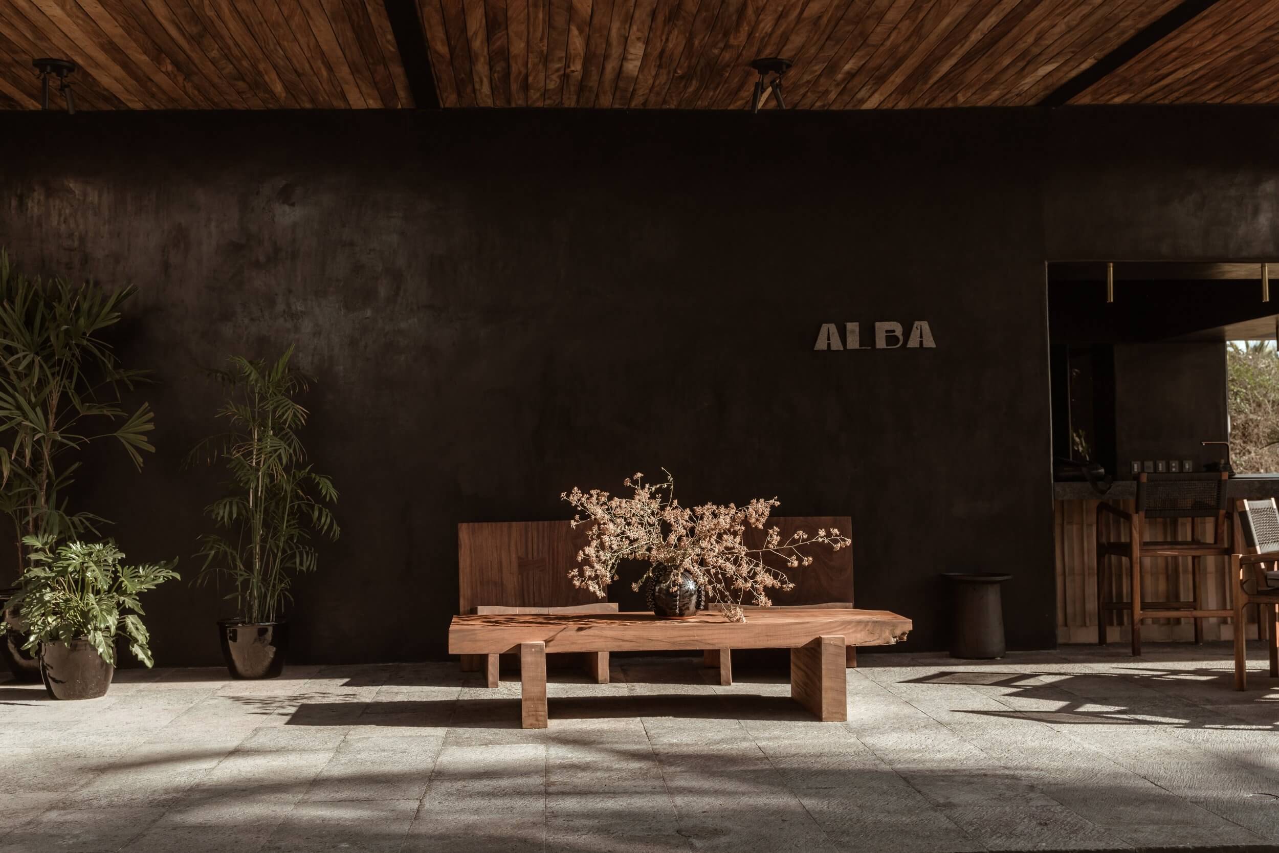 ALBA, Mexico — Minimalist wooden bench beneath dark concrete wall with dried botanical arrangement, natural light casting geometric shadows on stone courtyard.