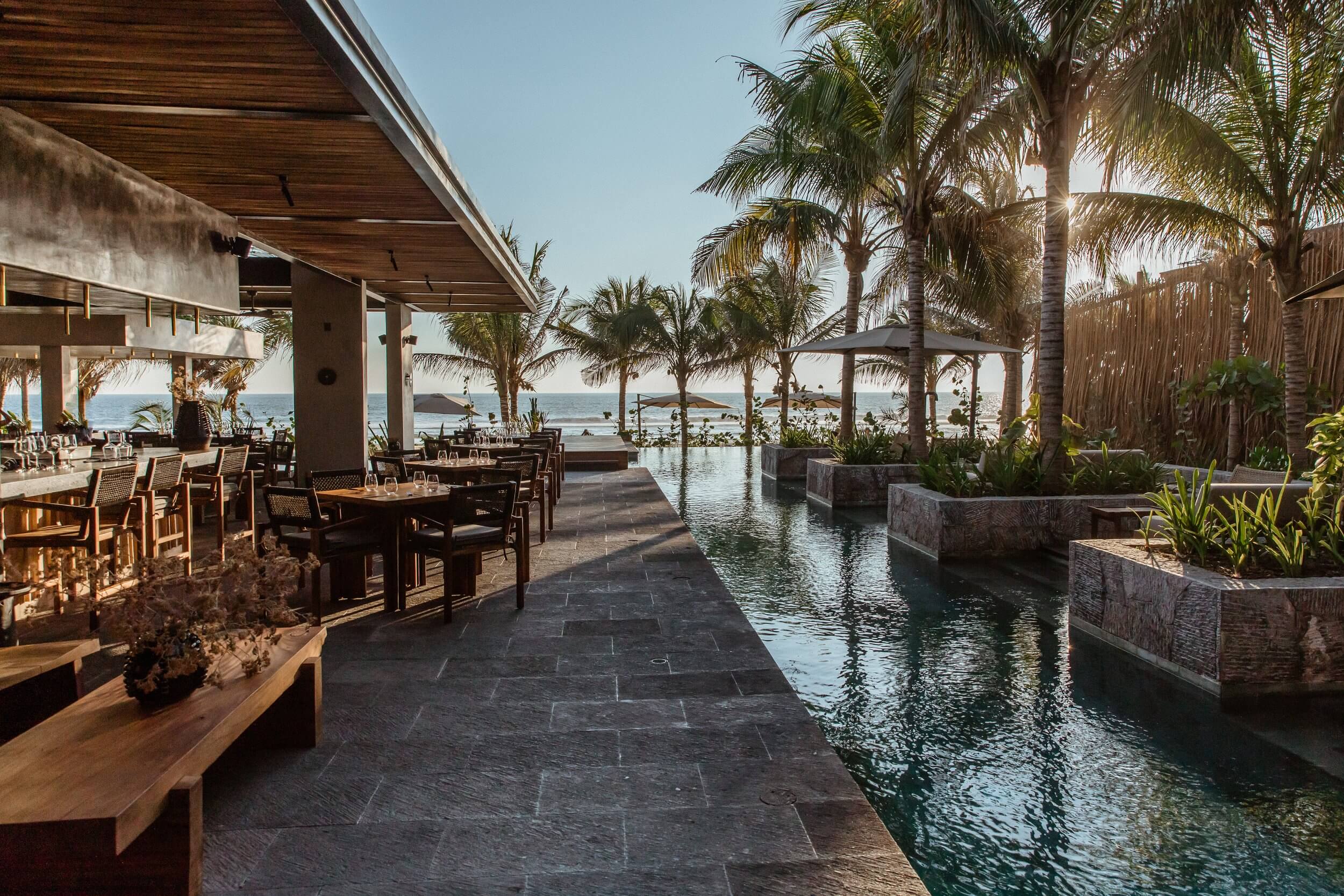 Hotelito MUSA, Mexico — Oceanfront dining pavilion with timber ceiling, slate flooring, and infinity pool reflecting palm trees and sea horizon at dusk.