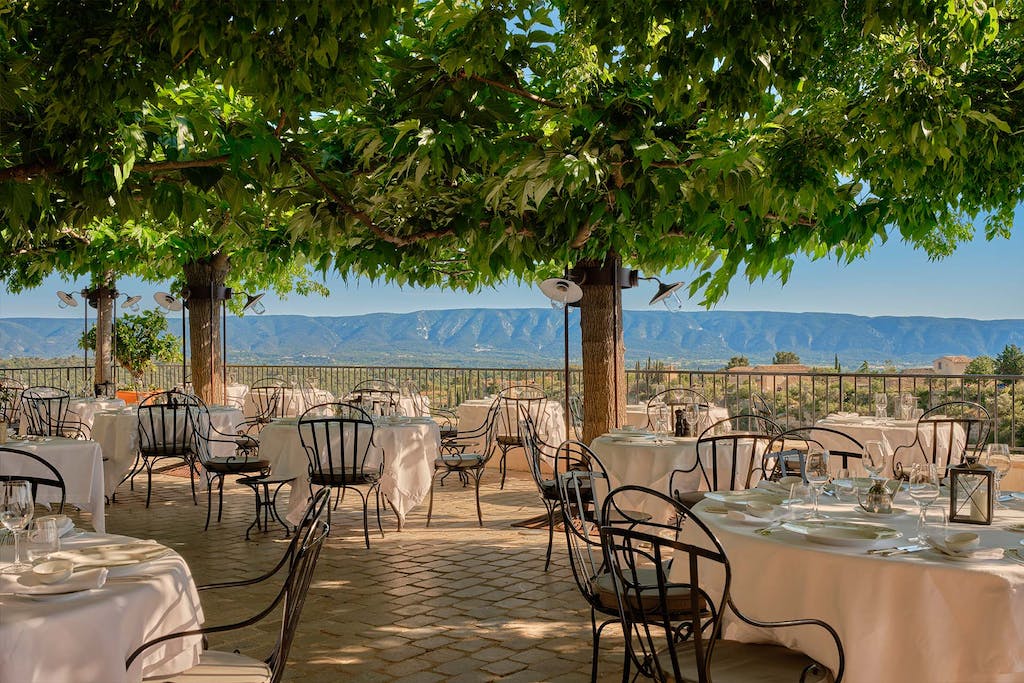 L’Orangerie — L'Orangerie, Europe — Shaded terrace dining with wrought-iron chairs, linen tablecloths, and panoramic valley views beneath mature trees.