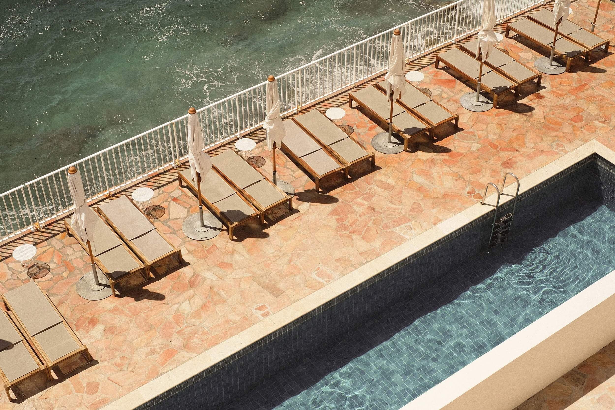 Les Roches Rouges, Europe — Sunlit terrace with teak loungers, cream umbrellas, and terracotta stone overlooking a dark-tiled pool and turquoise water beyond.