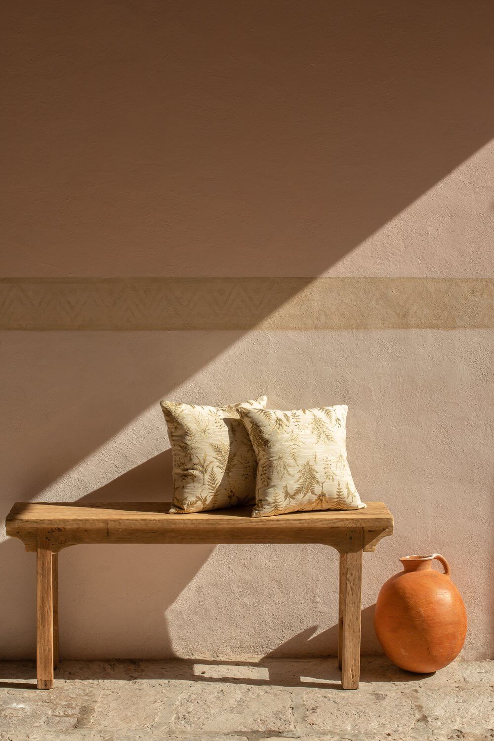 La Valise San Miguel, Mexico — Wooden bench with cream fern-print pillows beside terracotta vessel, sunlit minimalist adobe wall.