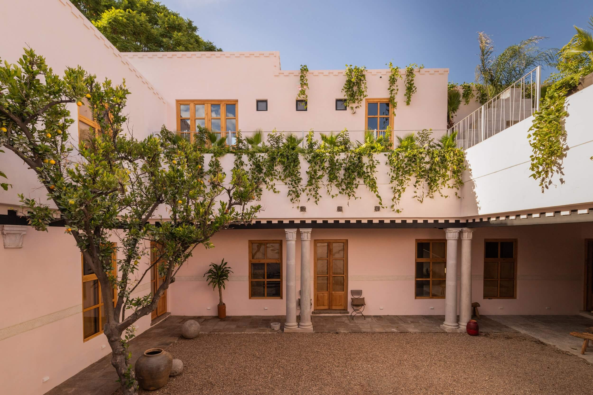 La Valise San Miguel, Mexico — Pink-washed courtyard with colonial columns, hanging vines, wooden doors, terracotta vessels, and upper-level windows under clear sky.