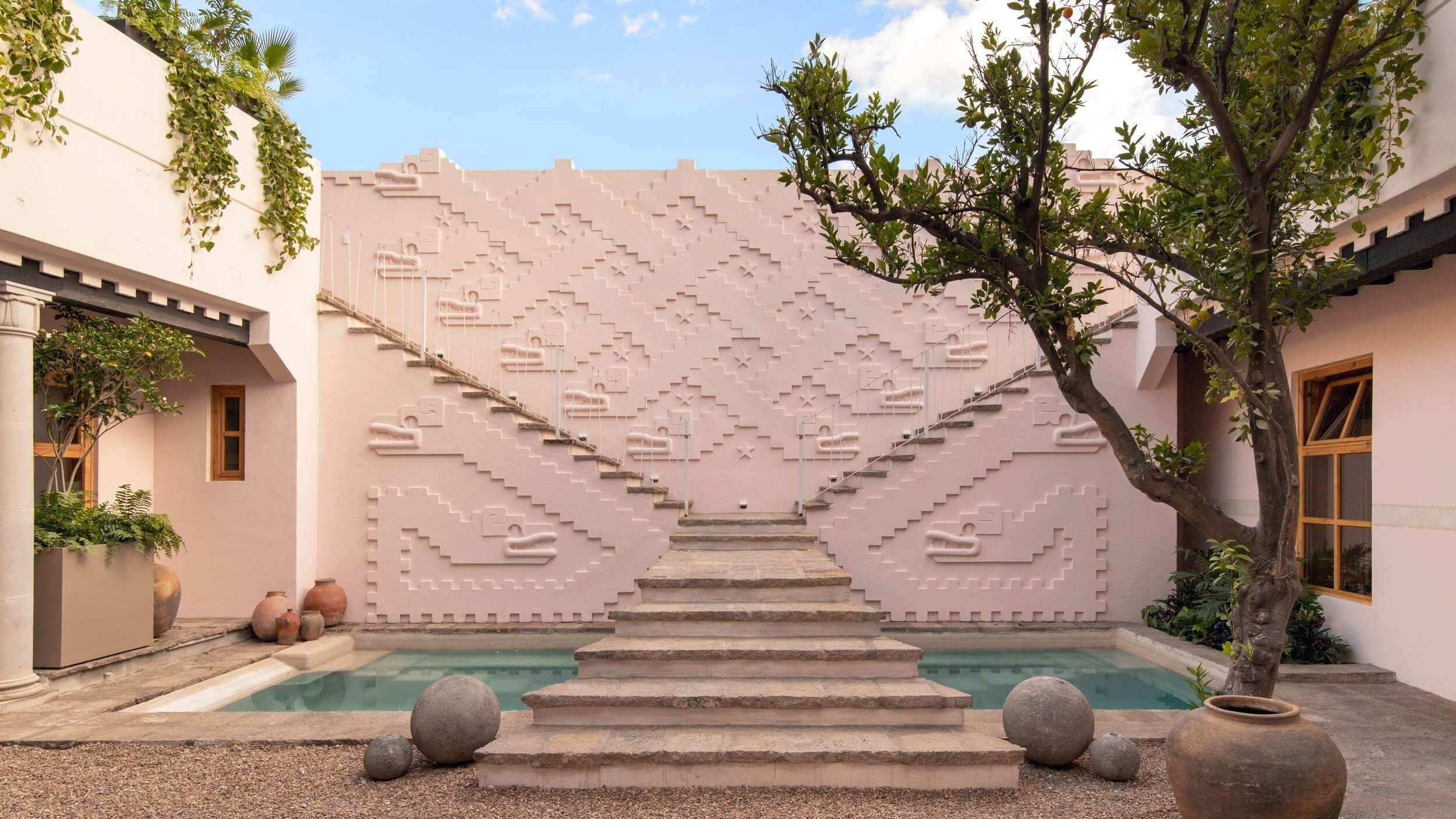 La Valise San Miguel, Mexico — Minimalist courtyard with geometric pink stucco wall, turquoise pool, stepped platform, mature tree, terracotta vessels, and sculptural stone spheres.