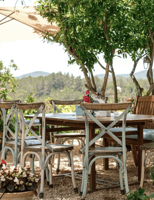 La Paloma, Balearic Islands — Weathered dining table and chairs beneath lush tree canopy overlooking forested hillside, Mediterranean outdoor living space.
