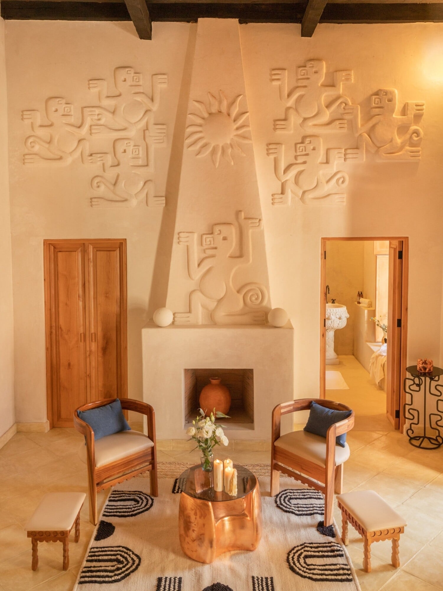 La Valise San Miguel, Mexico — Ochre stucco room with carved sun motif, wooden chairs, copper vessel, Aztec-inspired wall reliefs, and clay fireplace.