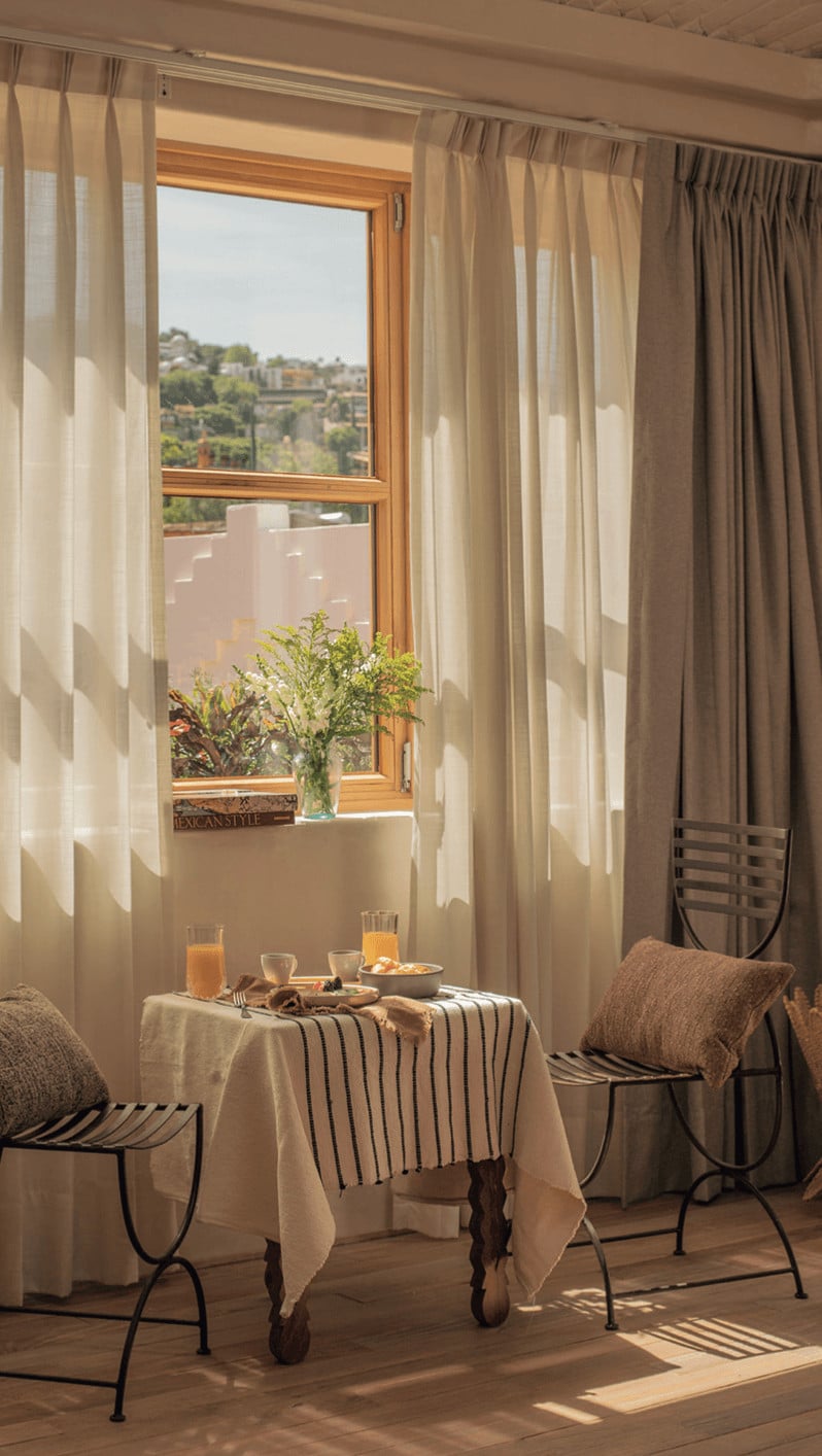 La Valise Breakfast, Mexico — Sunlit dining nook with sheer curtains, wrought-iron chairs, striped linen tablecloth, fresh flowers, and hillside vista through wooden-framed window.
