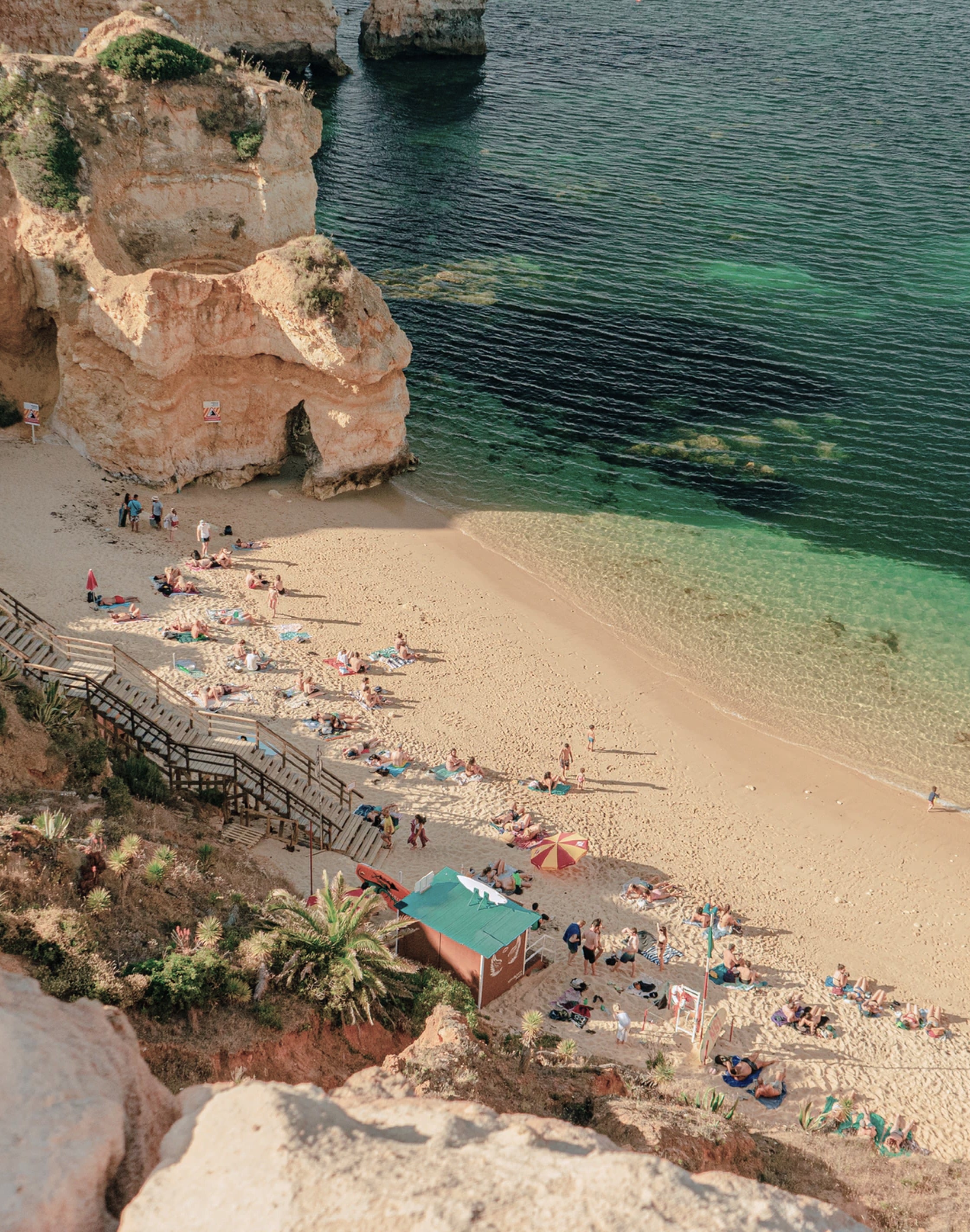 Casa Mãe, Europe — Terracotta cliffs frame sandy cove with turquoise waters, wooden beach access stairs, and sunbathing guests under umbrellas.