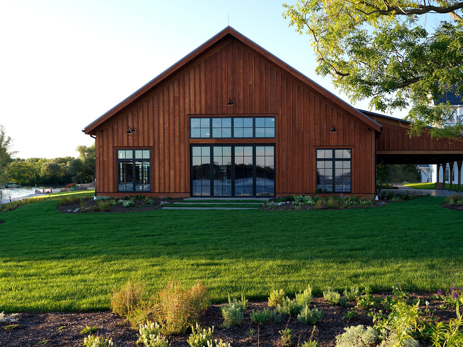 The Lake House, North America — Modern barn with dark wood siding, expansive grid windows, riverside setting, manicured lawn, contemporary hospitality architecture.