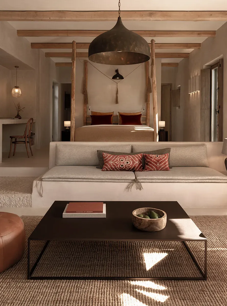 The Rooster, Europe — Minimalist villa bedroom with wooden four-poster bed, black pendant lamp, neutral linens, terracotta accents, and natural light across cream walls and natural fiber flooring.