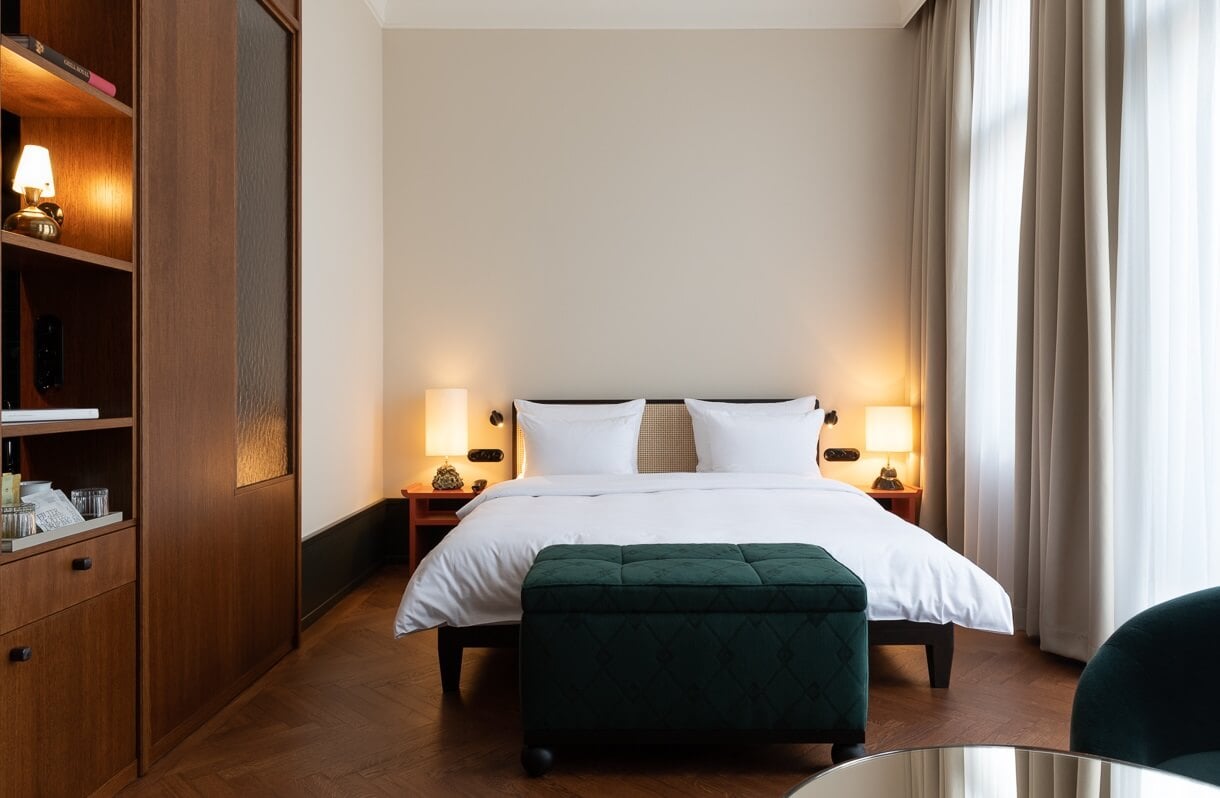 Château Royal, Europe — Luxury bedroom with warm wood cabinetry, crisp white bedding, deep green velvet bench, ambient brass lighting, and floor-to-ceiling curtains.