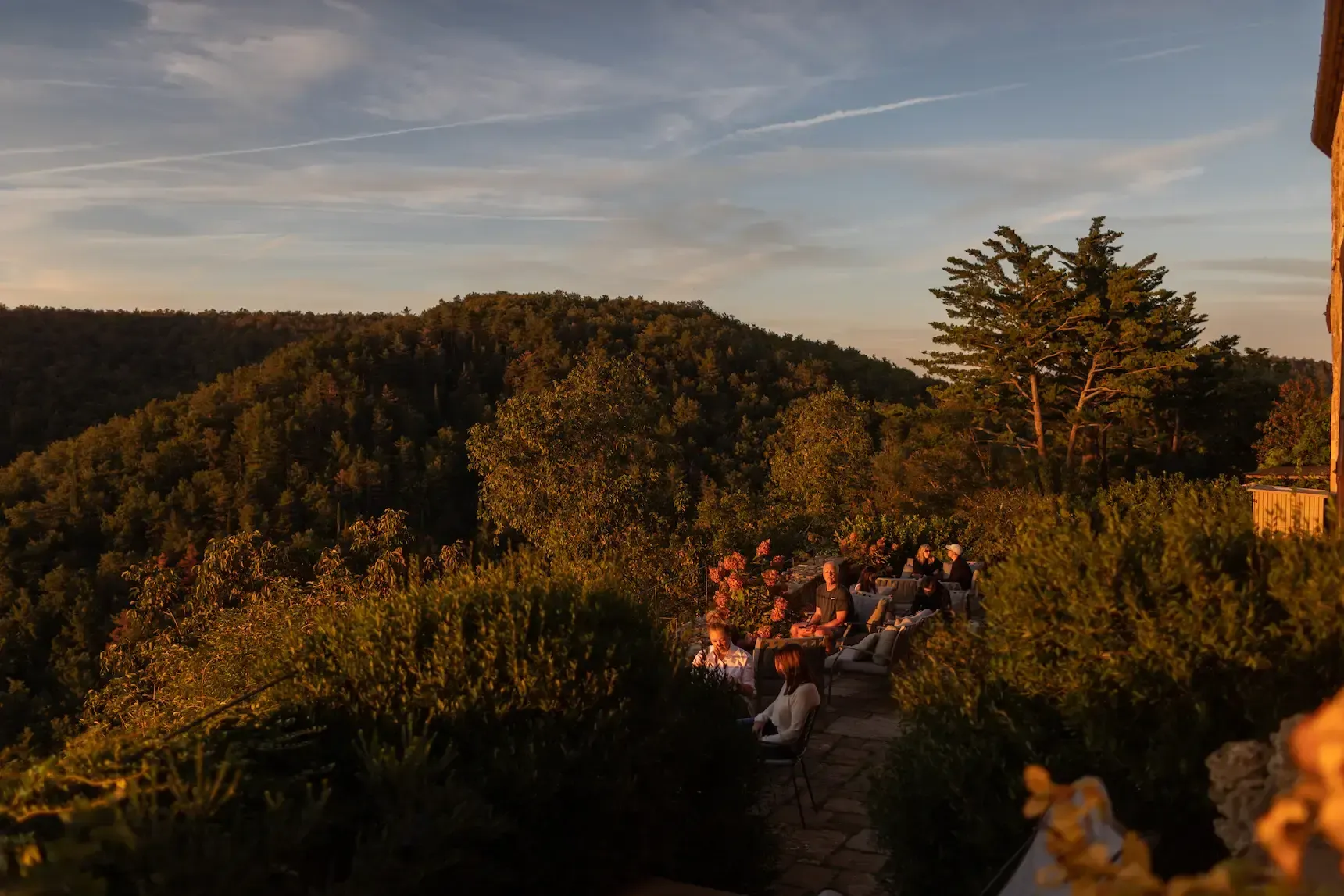 Lounge & Terrace Bar, Europe — guests gathered on hillside terrace at golden hour, surrounded by manicured hedges and forest landscape, warm ambient lighting.