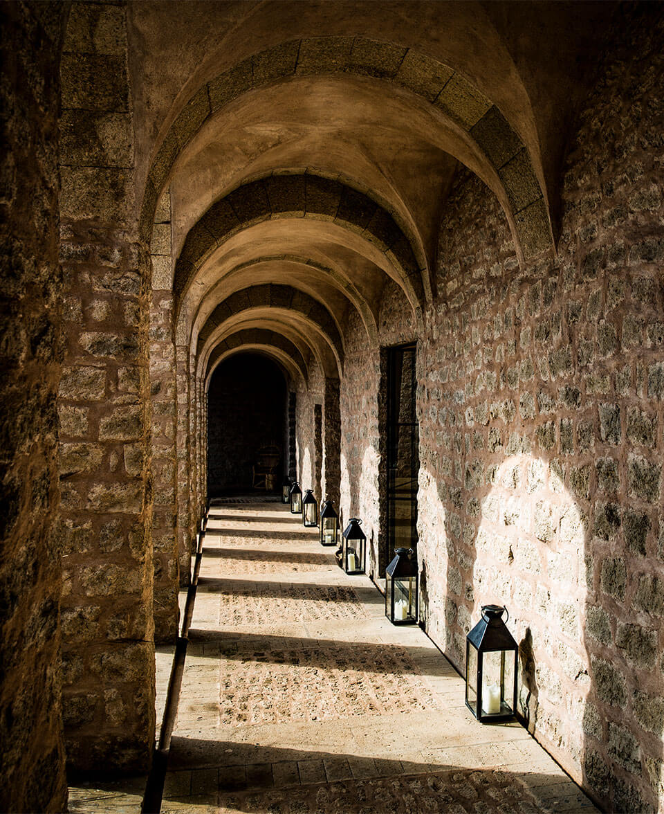 Arijiju Retreat, Africa — Stone barrel-vaulted corridor with arched ceilings, weathered walls, lantern sconces, and dappled sunlight casting geometric shadows on sandy floor.