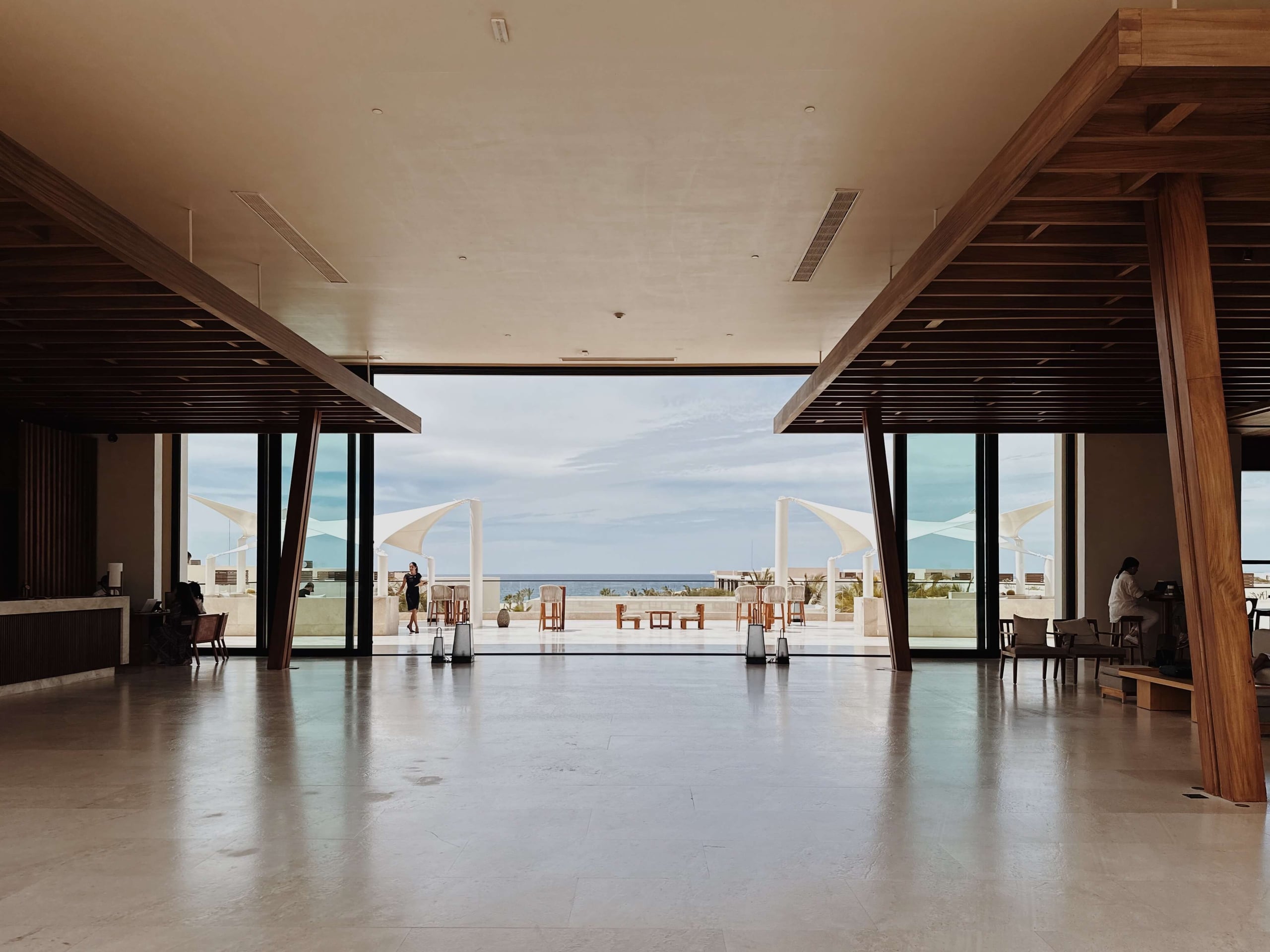Nobu Los Cabos, Mexico — Open-plan lobby with polished concrete floors, wooden ceiling beams, floor-to-ceiling windows framing sea views and sail canopies.