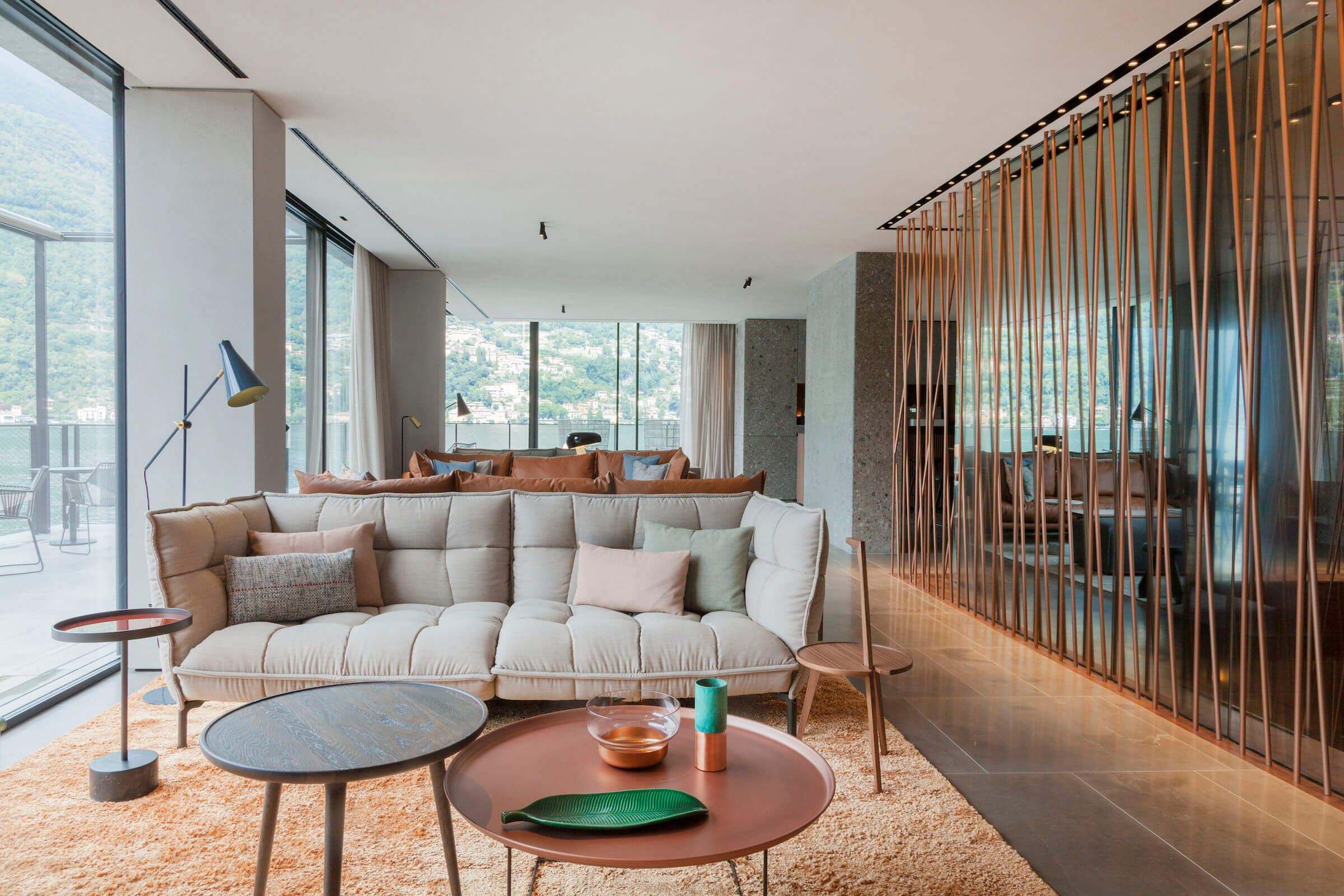il Sereno Lago di Como, Europe — Contemporary living room with cream quilted sofa, copper side tables, wooden partition screen, floor-to-ceiling windows overlooking alpine lake landscape.