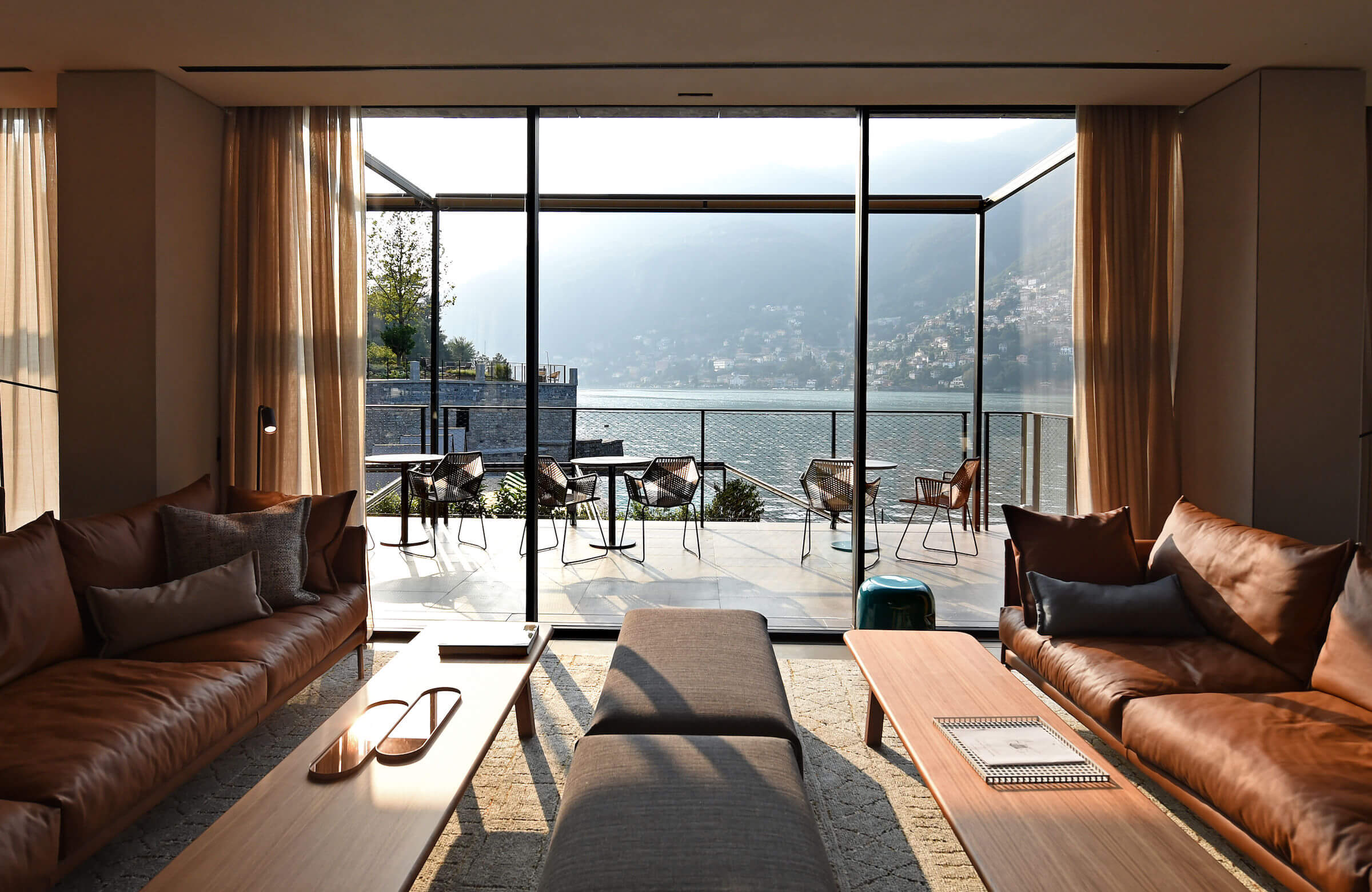 il Sereno Lago di Como, Europe — Lakeside lounge with cognac leather seating, floor-to-ceiling windows framing Alpine mountains, terrace with dining chairs overlooking misty water.