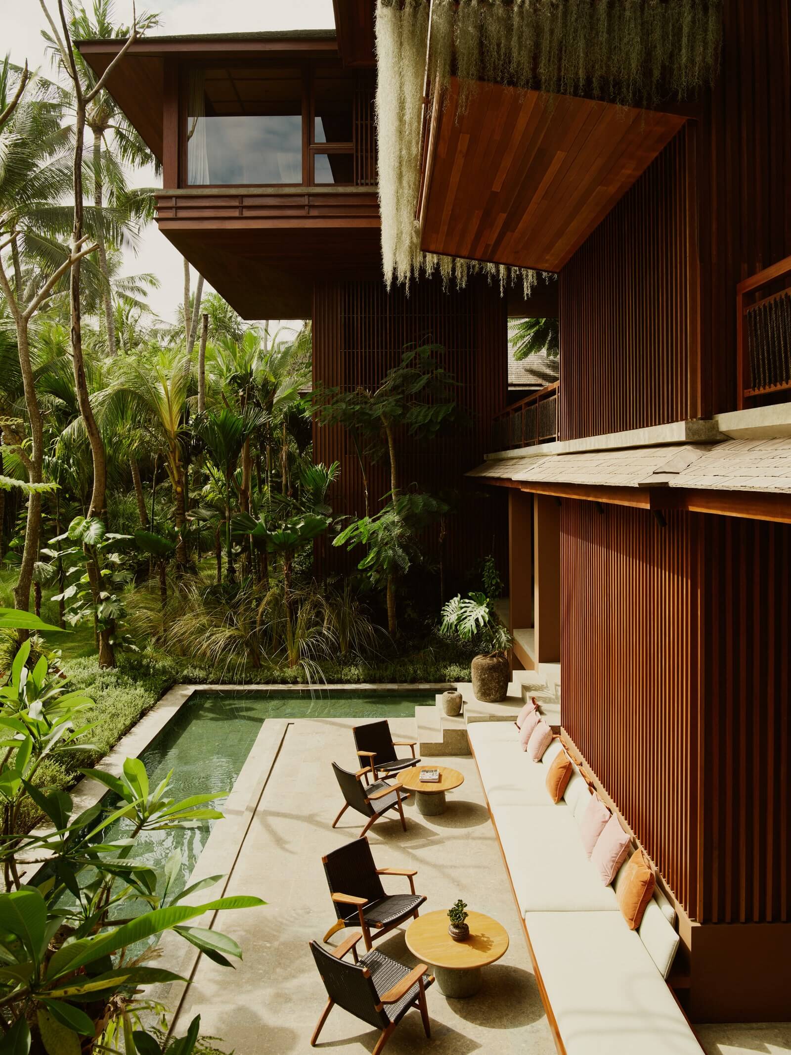 Lost Lindenberg, Asia — Tropical courtyard with wooden architecture, reflecting pool, modernist lounge chairs, lush vegetation, and natural light filtering through timber canopy.