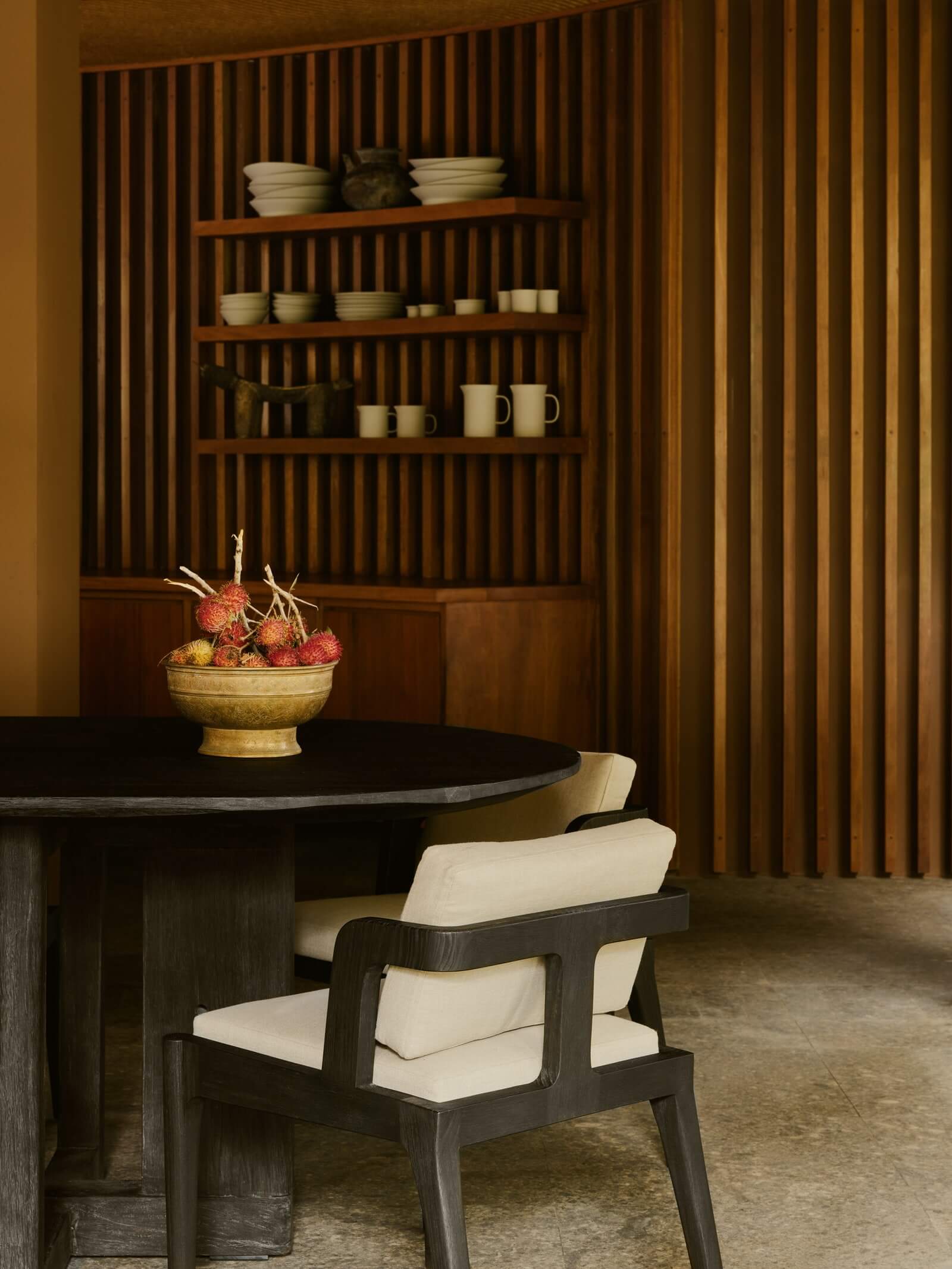 Lost Lindenberg, Asia — Minimalist dining space with black round table, dark wood chair with cream cushions, gold bowl of rambutans, vertical wooden slat wall, and open shelving displaying ceramic tableware.