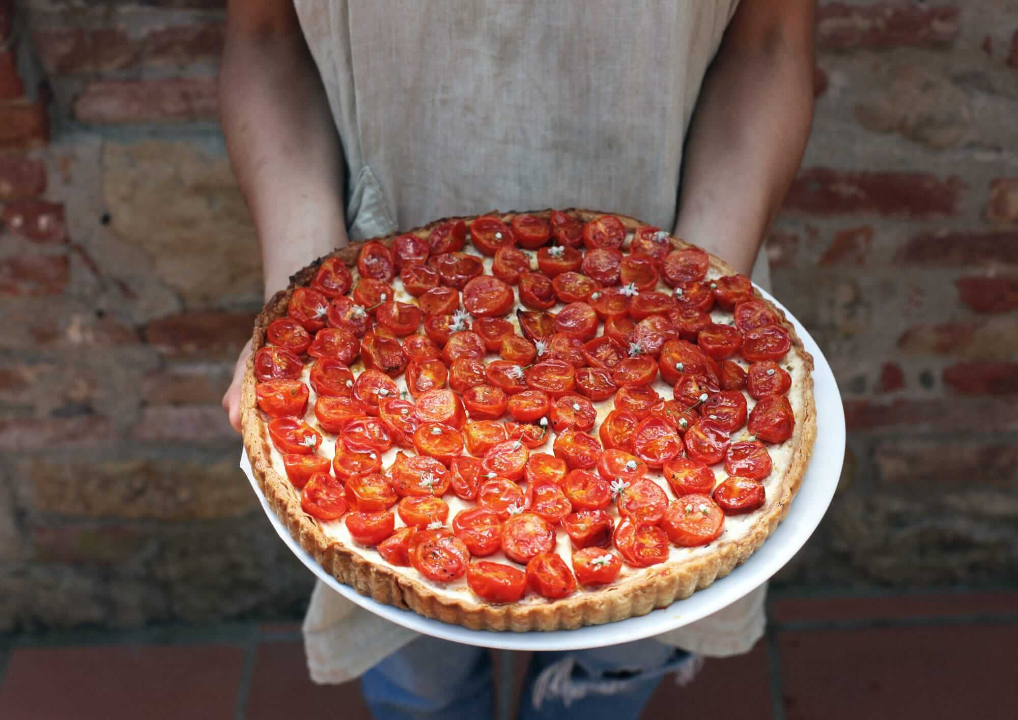 Villa Lena, Europe — Person holding freshly baked tomato tart with crisp pastry crust and roasted cherry tomatoes against rustic brick backdrop.