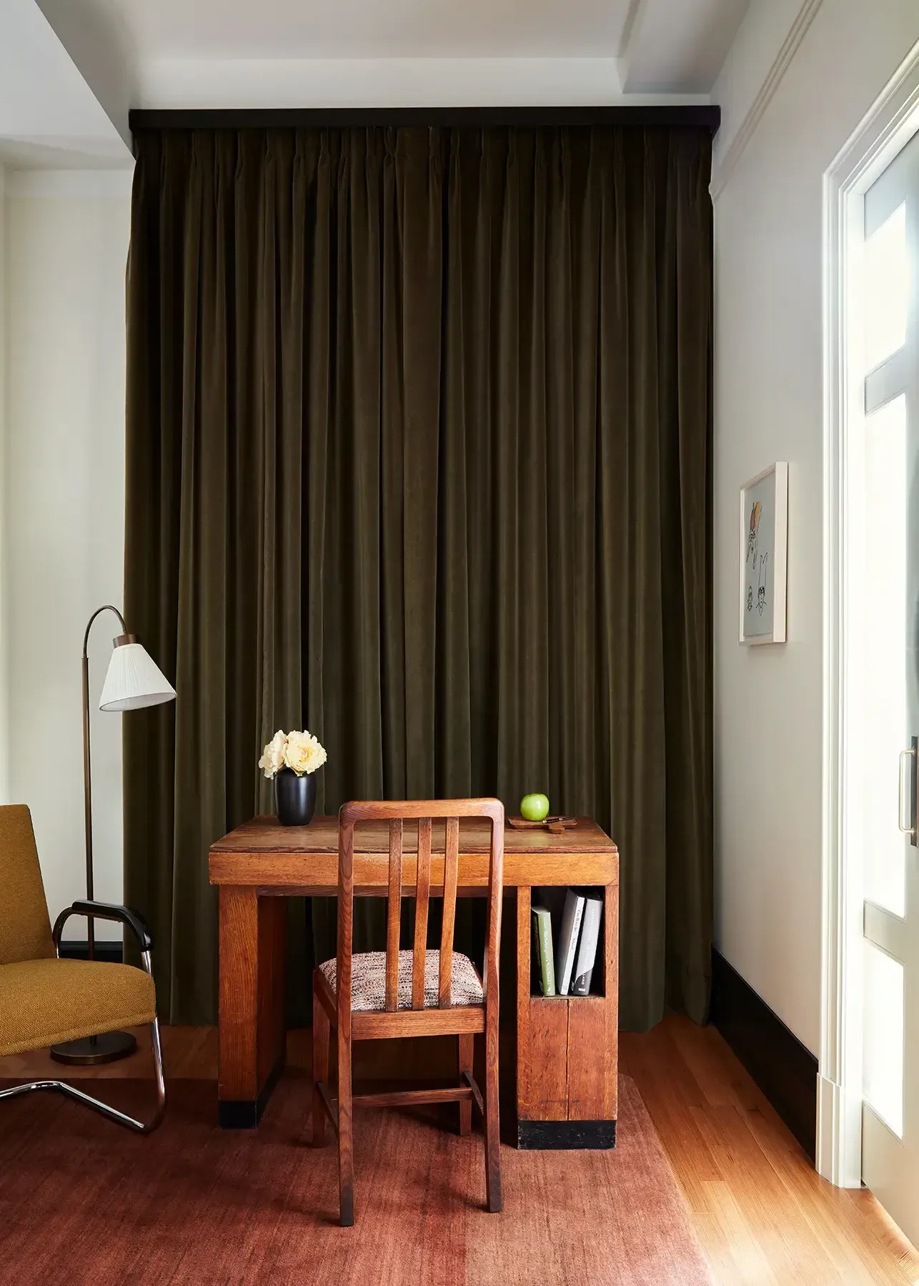 Nine Orchard, North America — Warm wood desk and chair beneath olive curtains, arc lamp and fresh flowers in modernist hotel workspace.