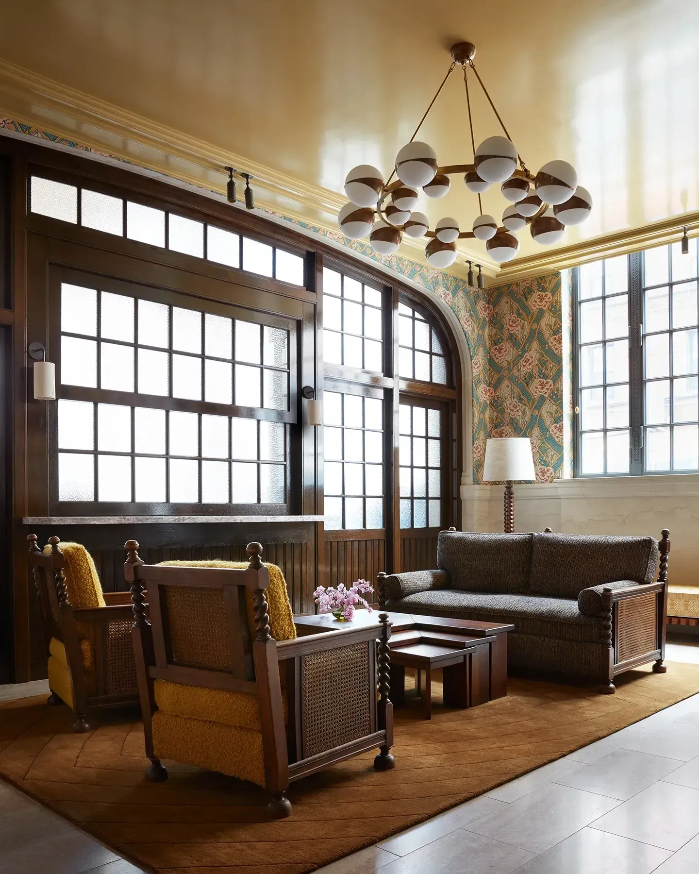 Nine Orchard, North America — Sunlit living room with brass pendant chandelier, dark wood-frame windows, mustard upholstered chairs, floral wallpaper, and modern furnishings.