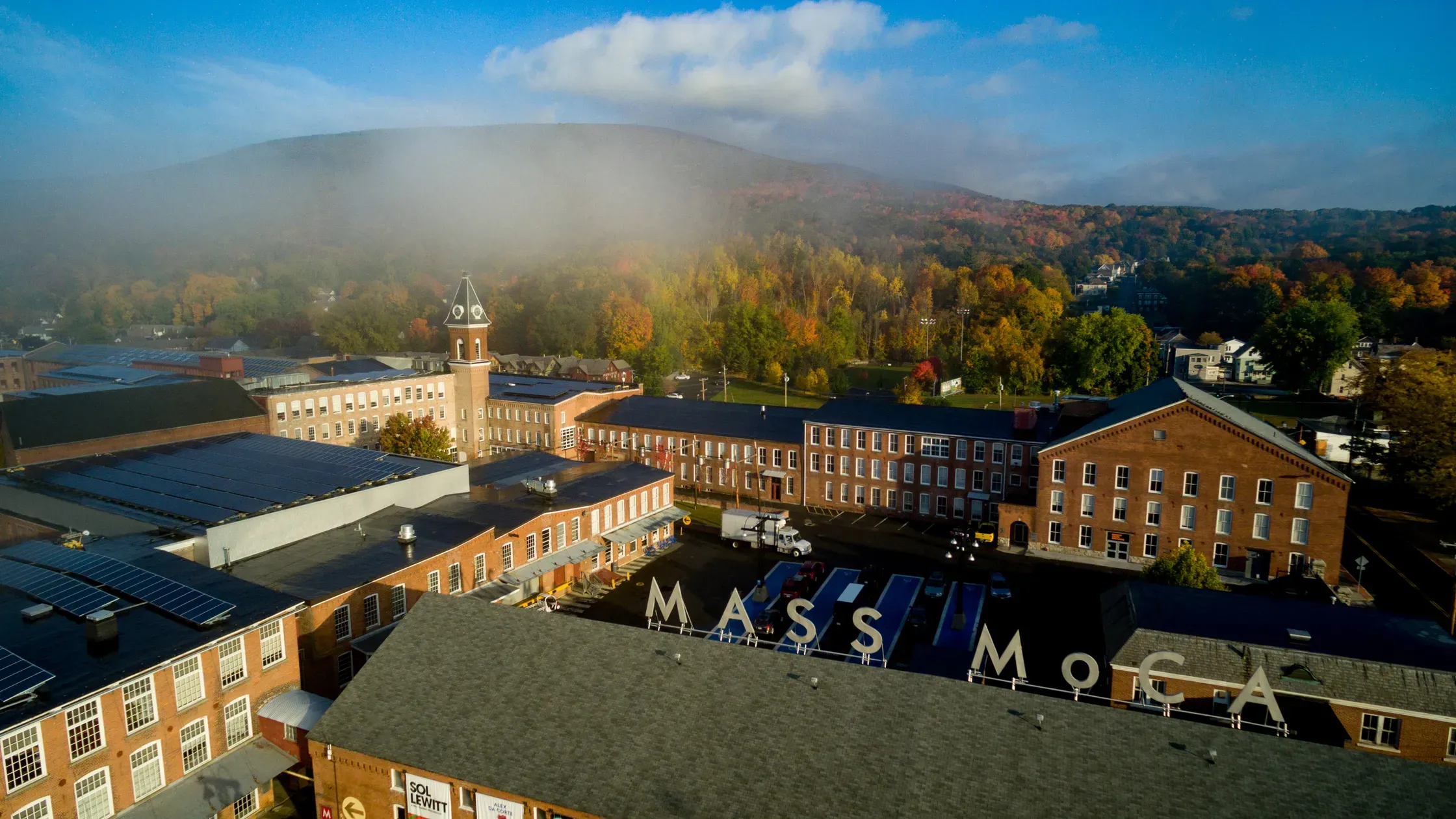 MASS MoCA, North Adams, Massachusetts — Autumn aerial view of brick industrial campus with clocktower, forested valley, and misty mountains under blue sky.