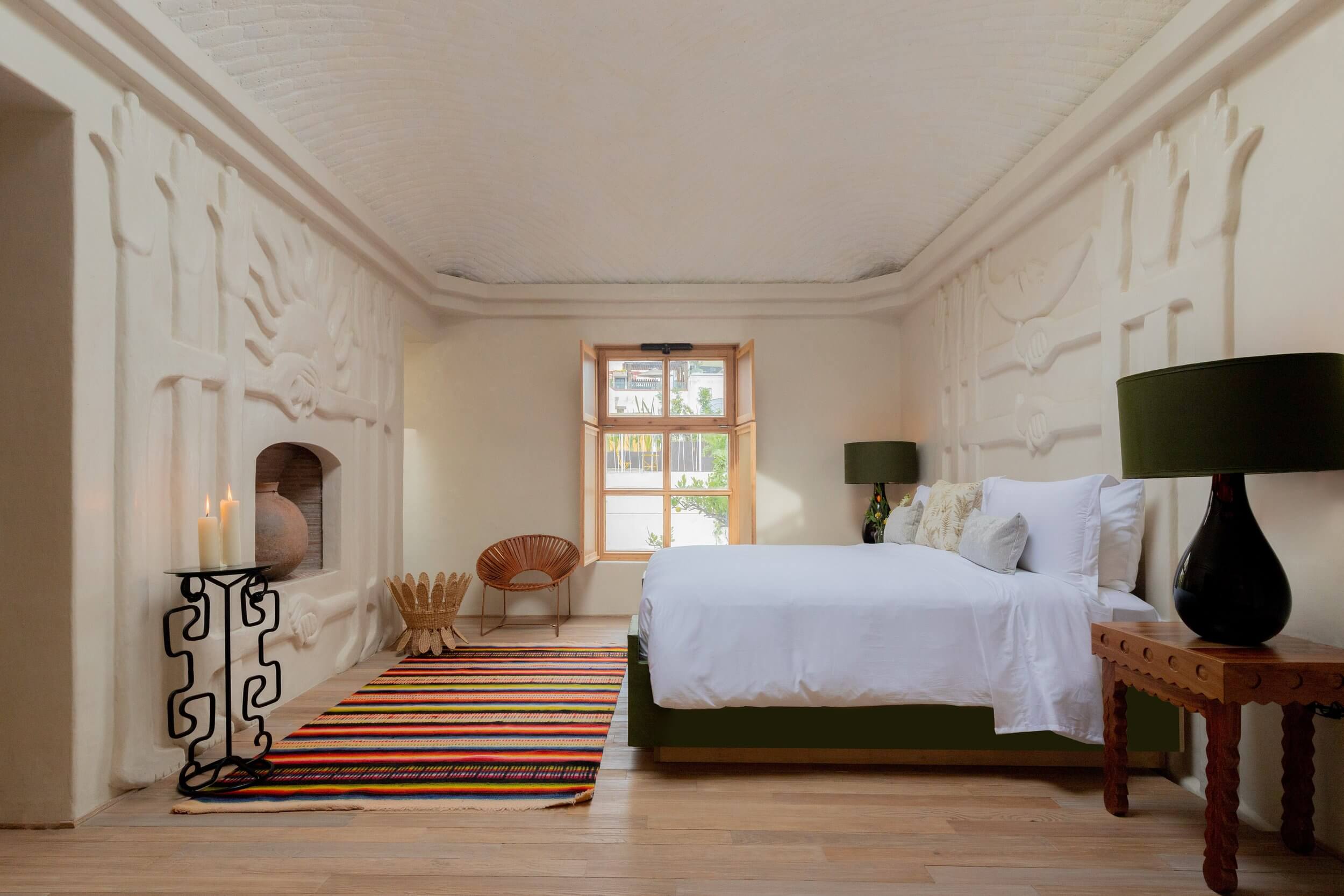 La Valise San Miguel, Mexico — Minimalist bedroom with cream plaster walls, wooden window frame, green upholstered bed, striped rug, and sculptural furnishings bathed in natural light.