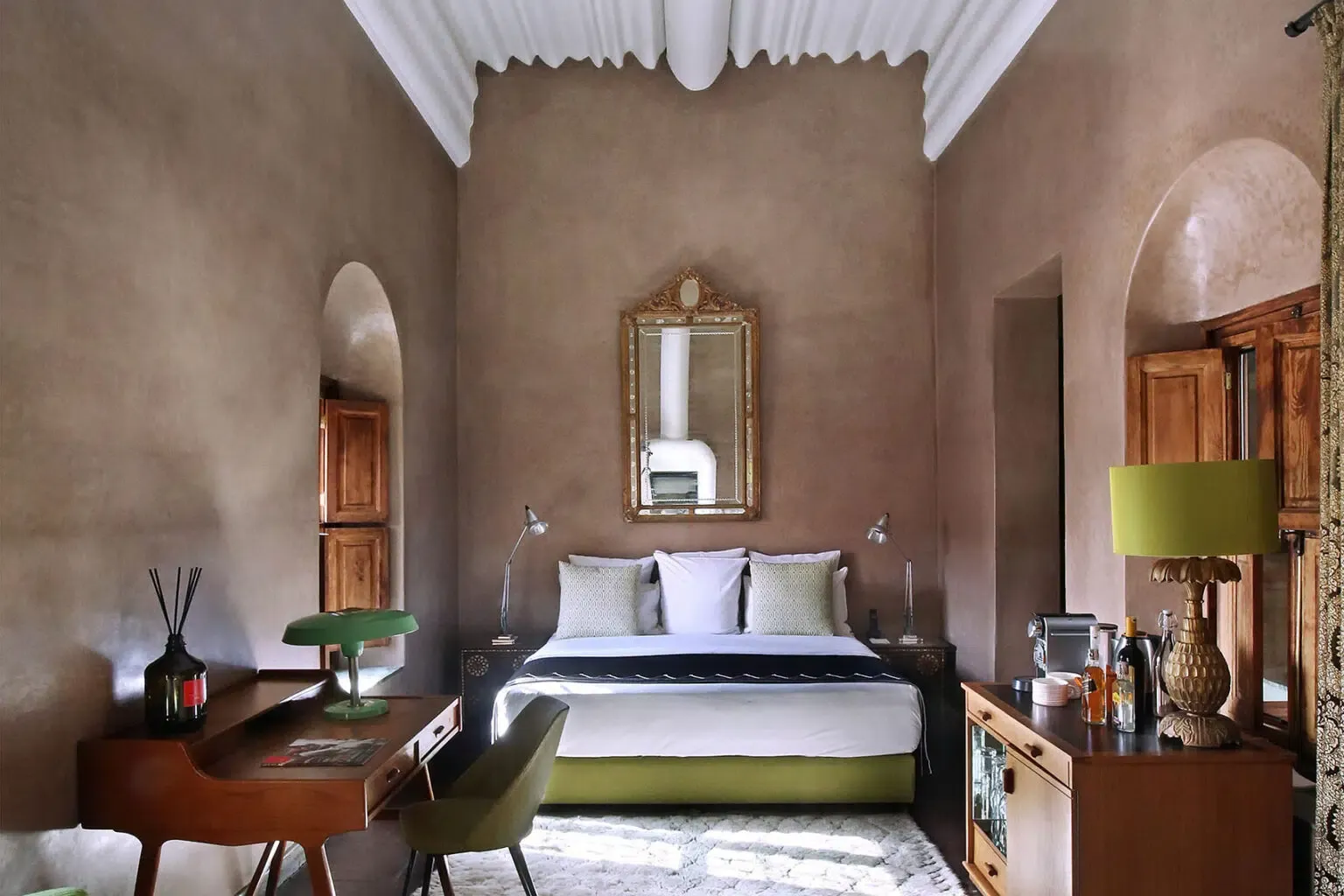 El Fenn, Marrakech, Morocco — Ochre bedroom with arched alcoves, carved wooden doors, gilt mirror, earth-toned walls, lime-green accent bench and bedding, brass lamp, white ceiling drapes.