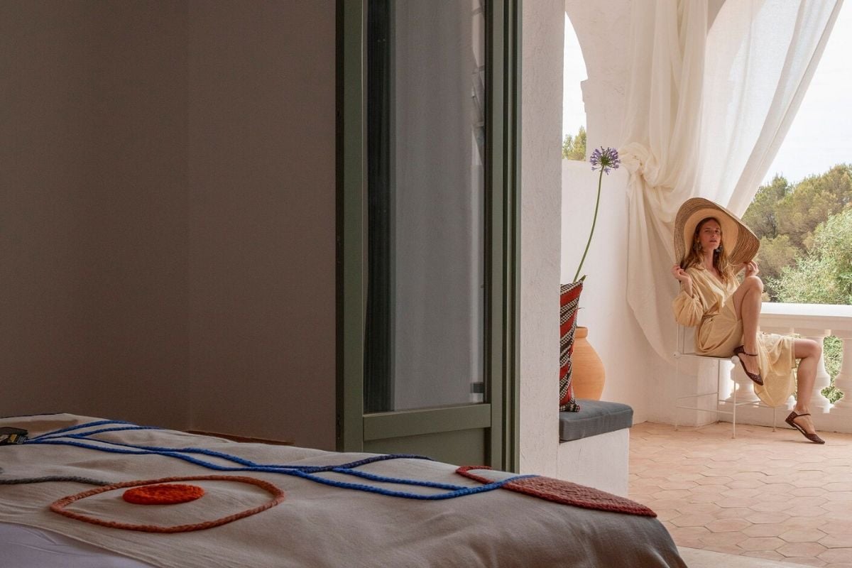 Menorca Experimental, Balearic Islands, Spain — Sunlit bedroom with sculptural rope installation, terracotta pottery, sheer curtains, woman in linen hat gazing toward Mediterranean garden vista.