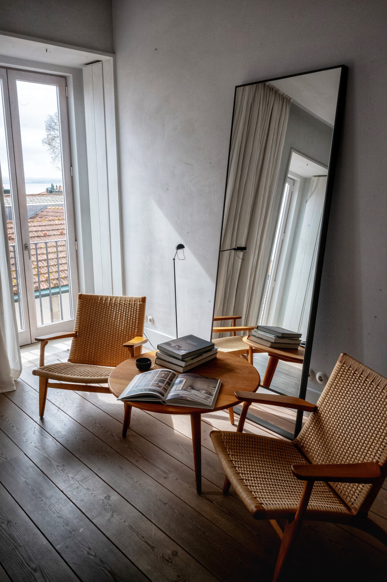 Santa Clara 1728, Portugal — Midcentury lounge chairs with woven seats frame a round wooden table beside a floor mirror, overlooking coastal rooflines through glass doors.