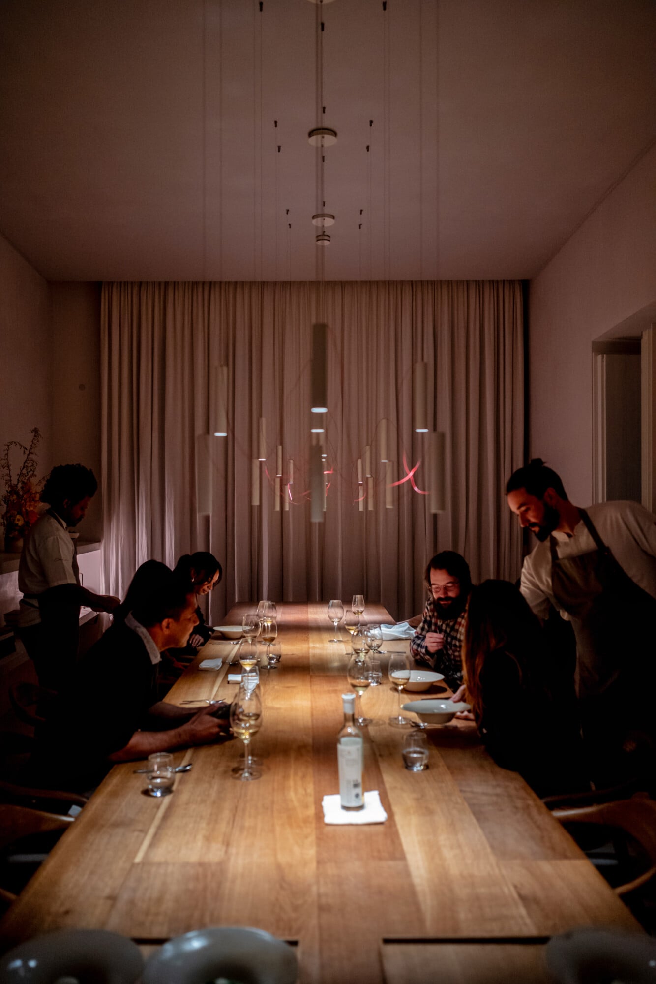 Santa Clara 1728, Portugal — Minimalist dining room with wooden table, neon signage, pendant lights, floor-to-ceiling curtains, and warm ambient lighting.