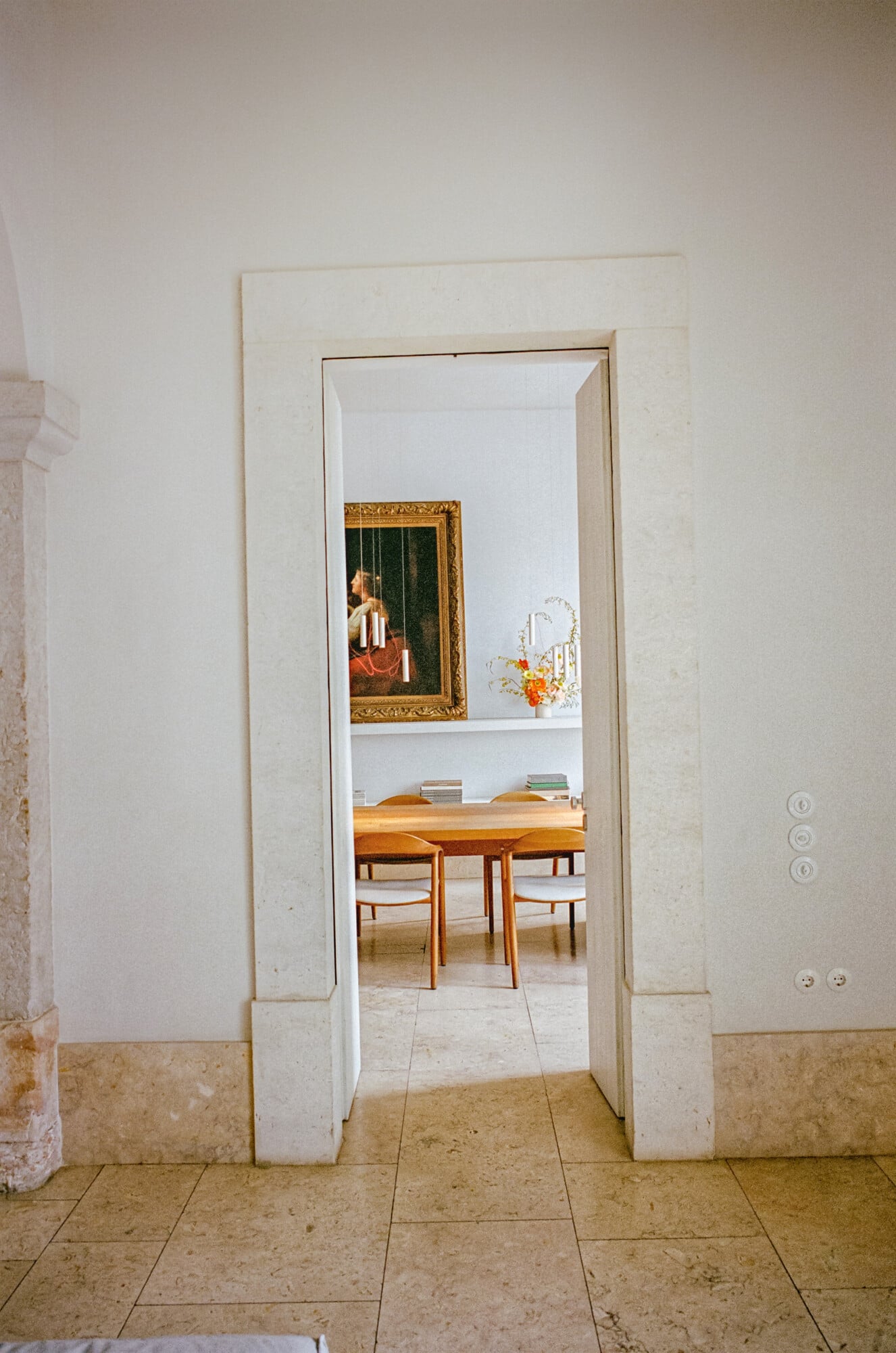 Santa Clara 1728, Portugal — Minimalist doorway with travertine frame opening onto mid-century dining room with wooden table, gilt-framed painting, and white walls.