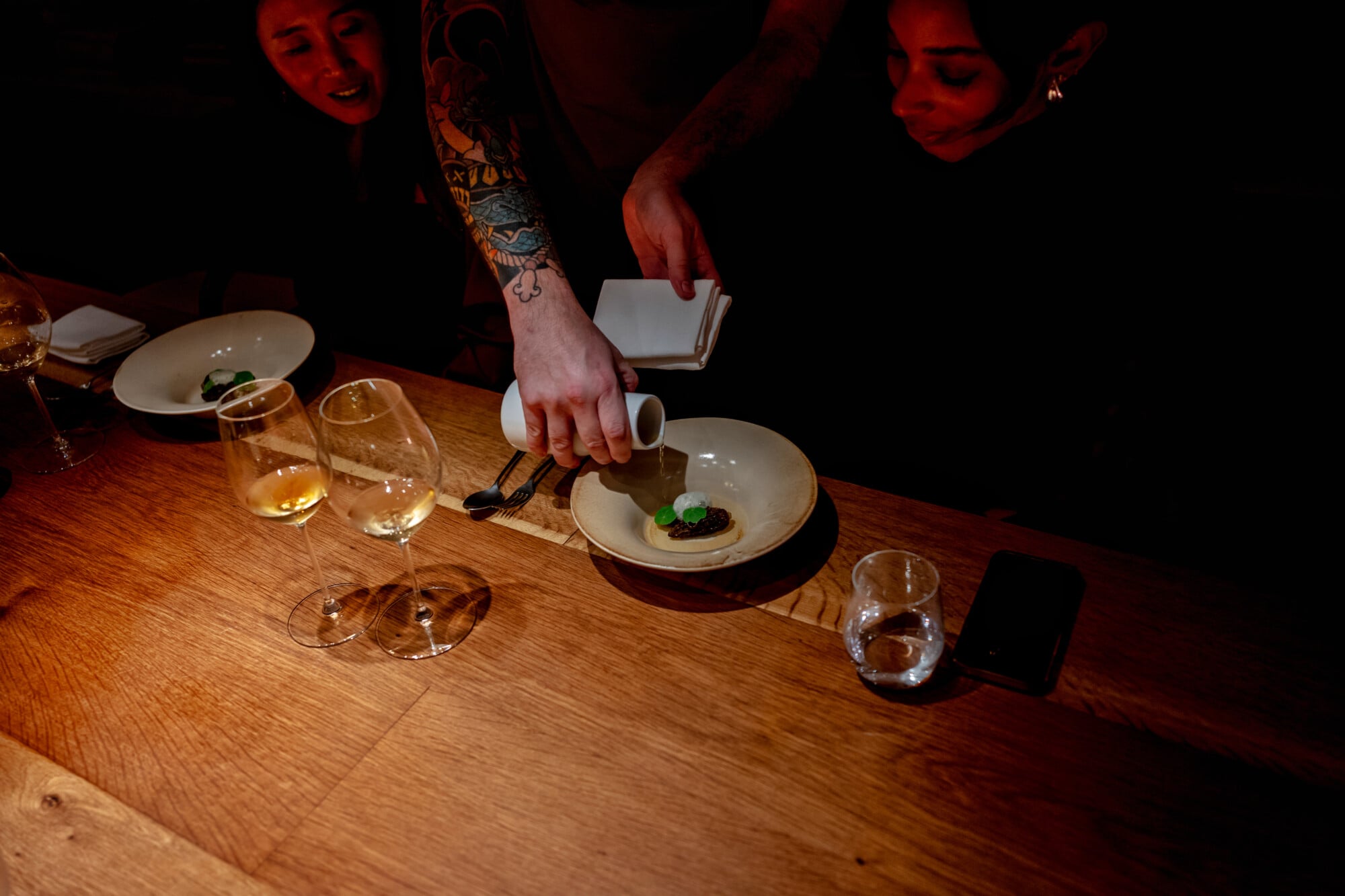 Santa Clara 1728, Portugal — Chef's hands plating artisanal dish with green microgreens at intimate wooden table with wine glasses and amber lighting.