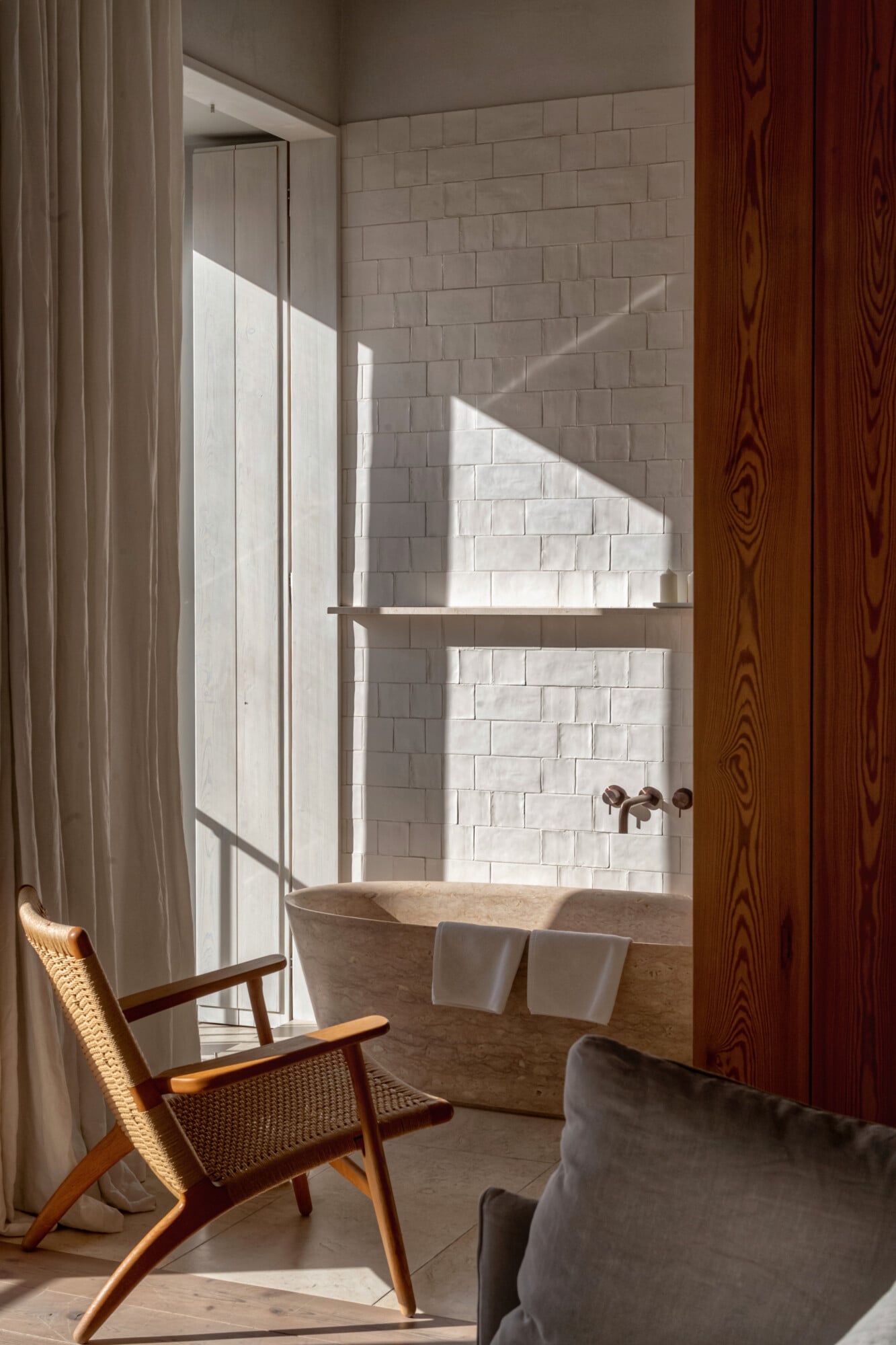Santa Clara 1728, Portugal — Sunlit bathroom with sculptural stone bathtub, white subway tile walls, mid-century modern chair, and natural light through glass partition.