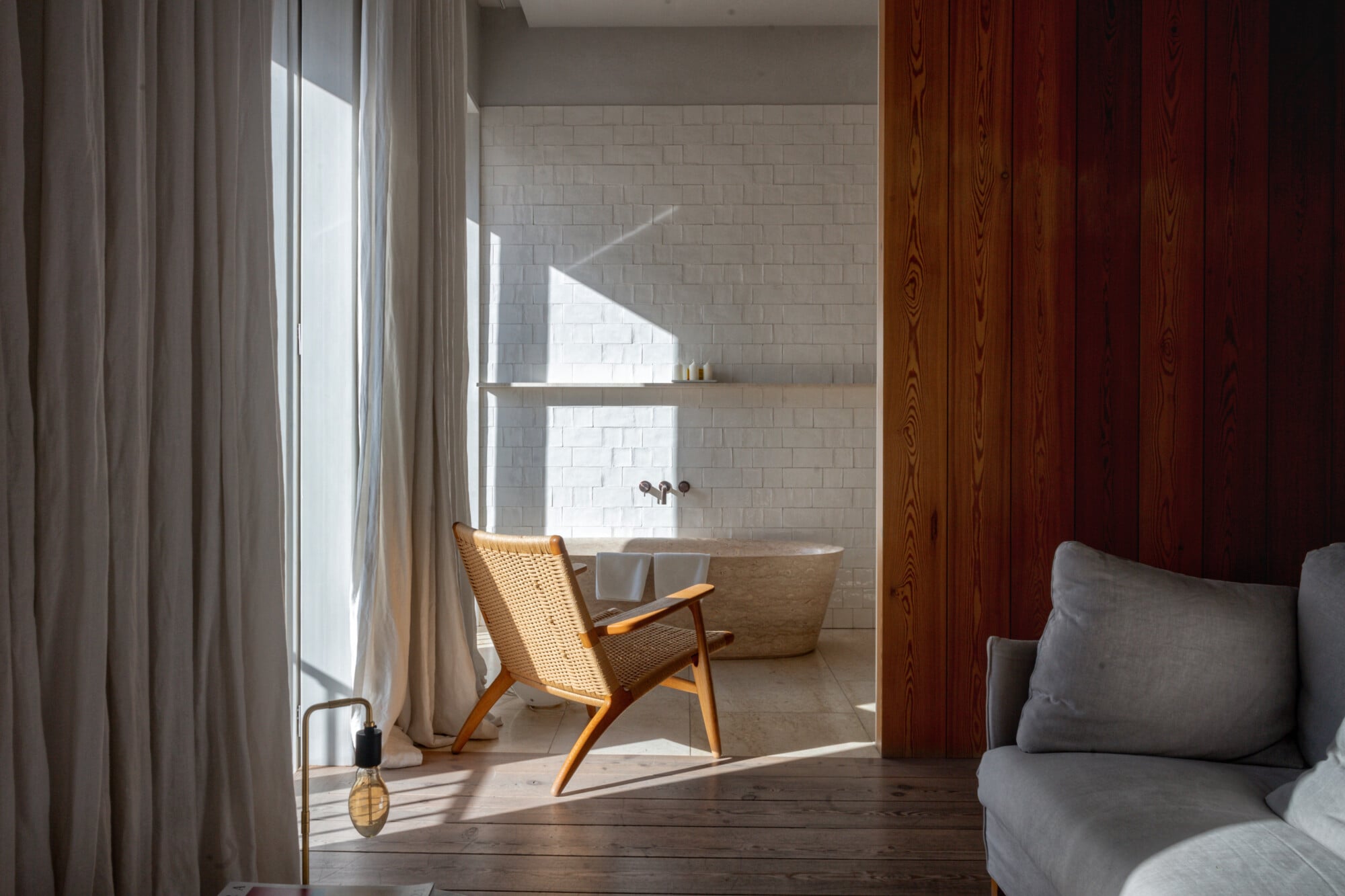 Santa Clara 1728, Portugal — Mid-century modern chair beside white subway-tiled bathroom with soaking tub, natural light through linen curtains, warm wood paneling, minimalist luxury spa aesthetic.